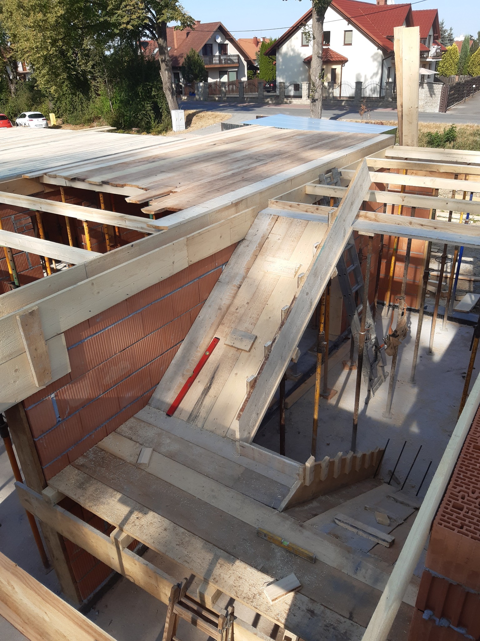 Budowa domu: Ściany z czerwonej cegły, drewniana konstrukcja stropu z deskowaniem, podpory stalowe, poziomica czerwona i żółta, widok z góry na plac budowy w słoneczny dzień.