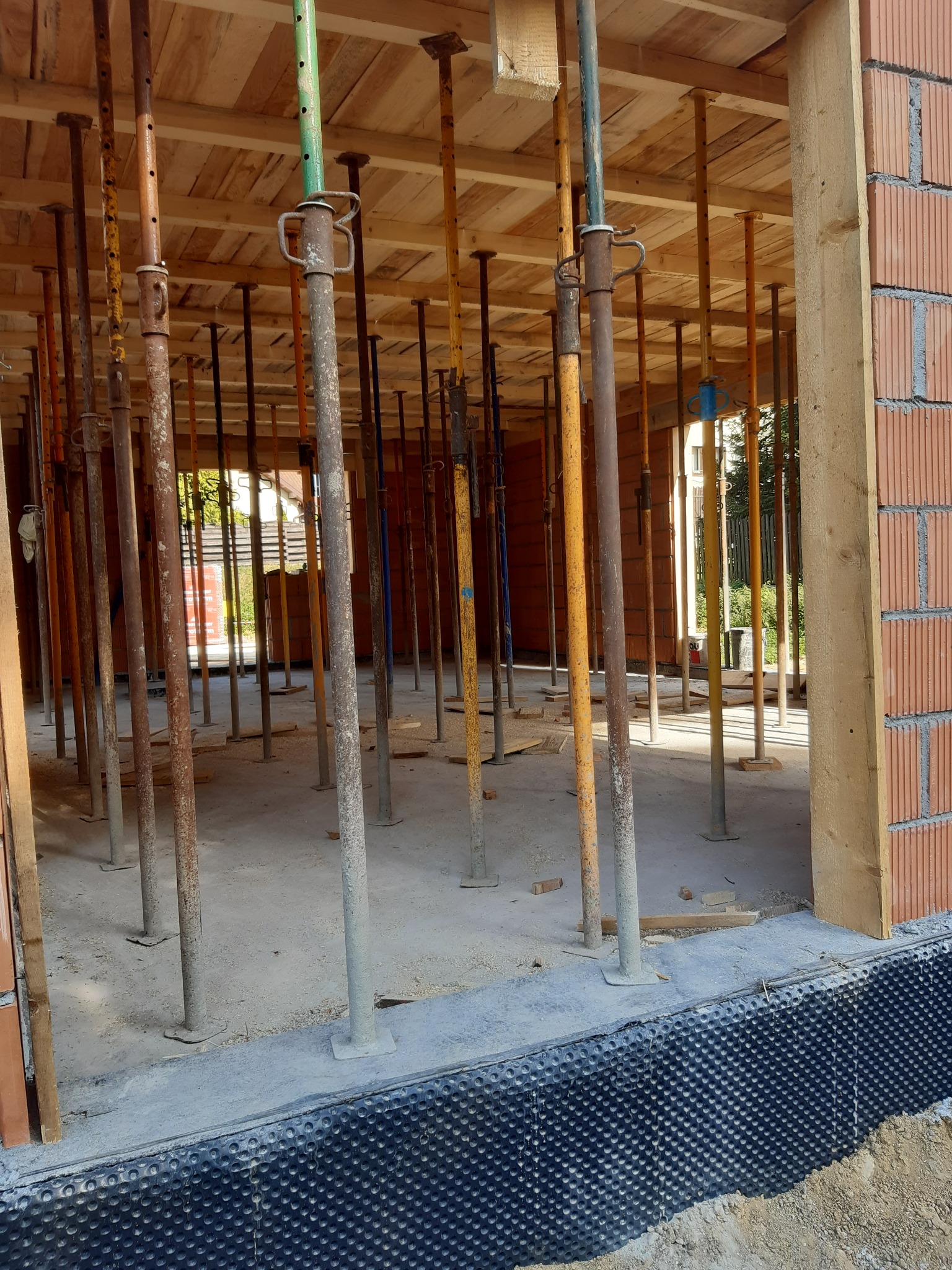 Wnętrze powstającego budynku z widocznymi podporami stropowymi, ceglanymi ścianami oraz izolacją przeciwwilgociową fundamentów w postaci folii kubełkowej.