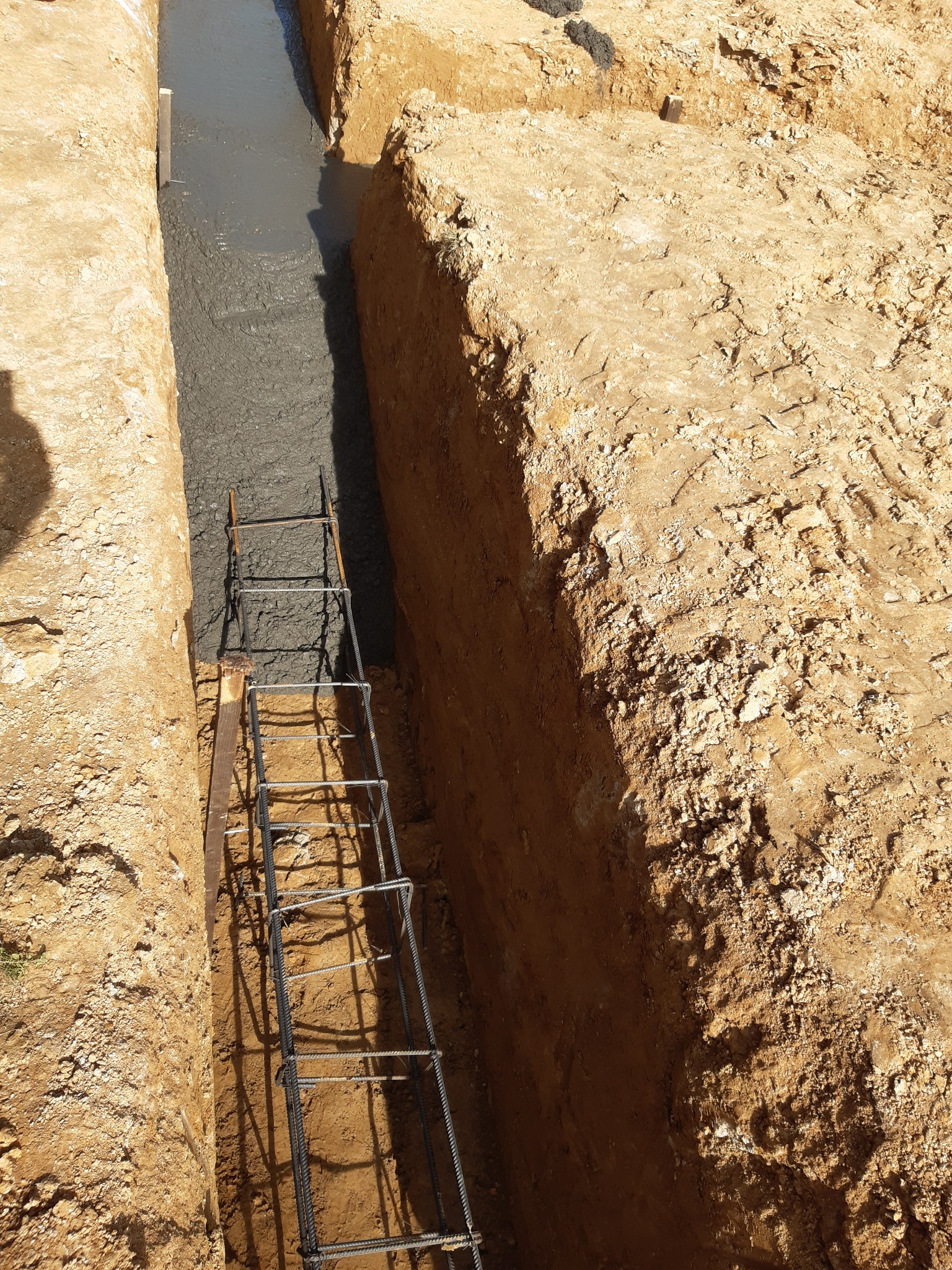 Wykop fundamentowy z zalewanym betonem i zbrojeniem stalowym, widok z góry na wąski wykop w ziemi.