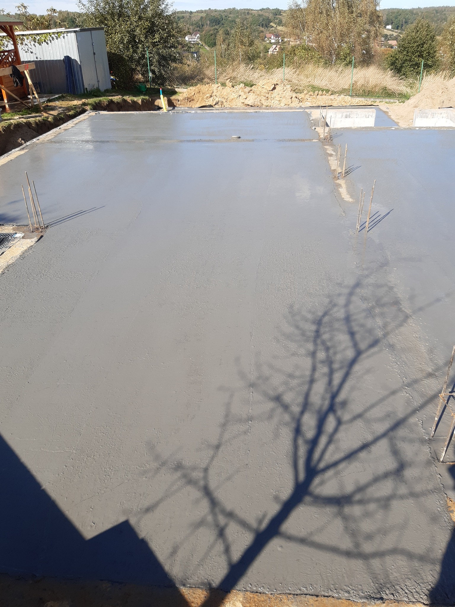 Świeżo wylana, gładka płyta betonowa fundamentowa z wystającymi prętami zbrojeniowymi, cień drzewa na powierzchni, widok z góry na plac budowy w słoneczny dzień.