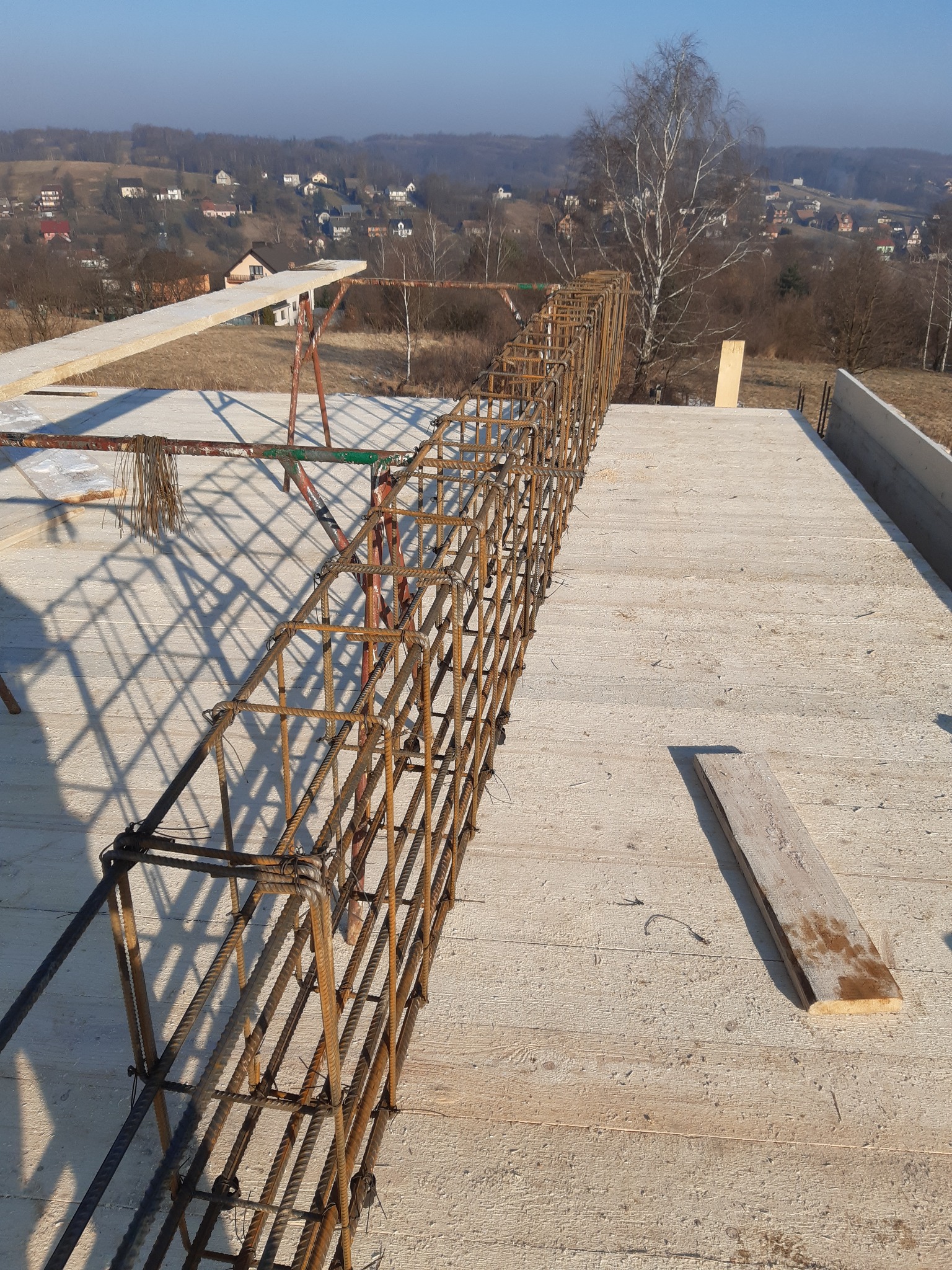 Zbrojenie fundamentu w trakcie budowy domu, widok z góry na betonową płytę z wystającymi prętami zbrojeniowymi, w tle zabudowa jednorodzinna na wzgórzu w słoneczny dzień.