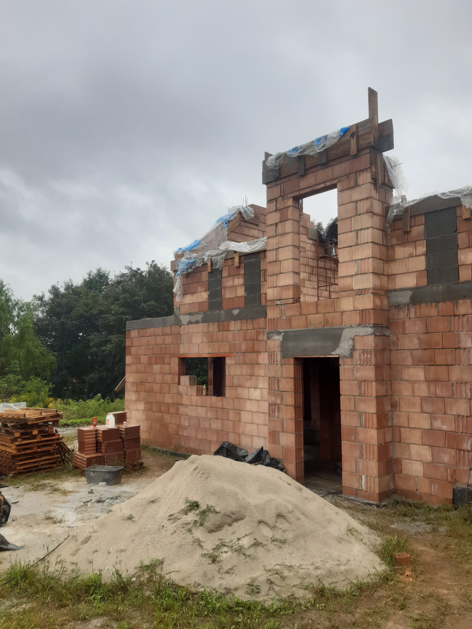 Wznoszenie ścian domu z czerwonej ceramiki poryzowanej; widoczny stos desek, zapas cegieł i piasek na placu budowy w pochmurny dzień.