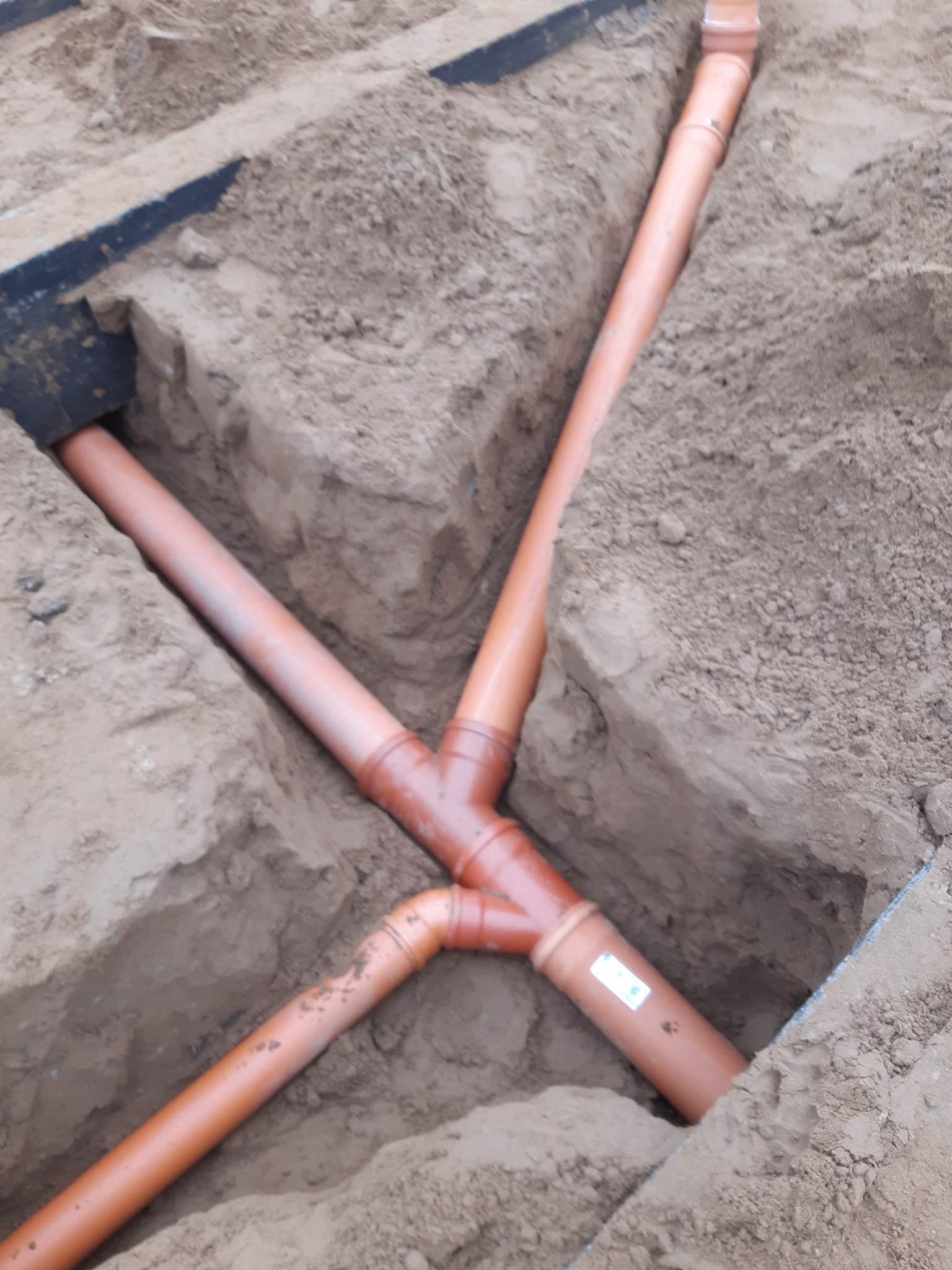 Pomarańczowe rury kanalizacyjne ułożone w wykopie w ziemi, z widocznymi złączkami i etykietą na jednej z rur.