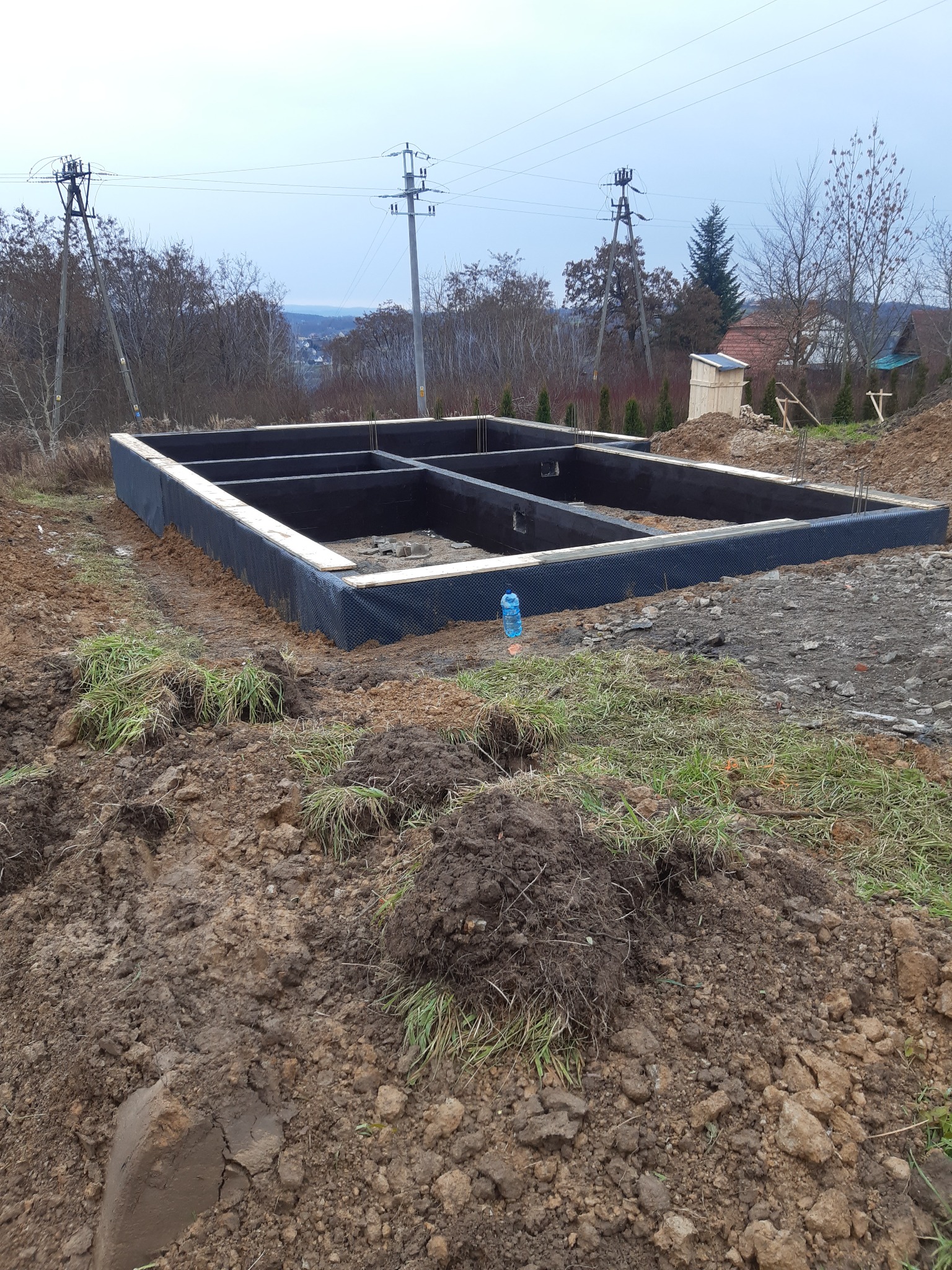 Wylane fundamenty domu jednorodzinnego z czarną izolacją pionową, widok z góry na wykop z wystającą butelką wody mineralnej.