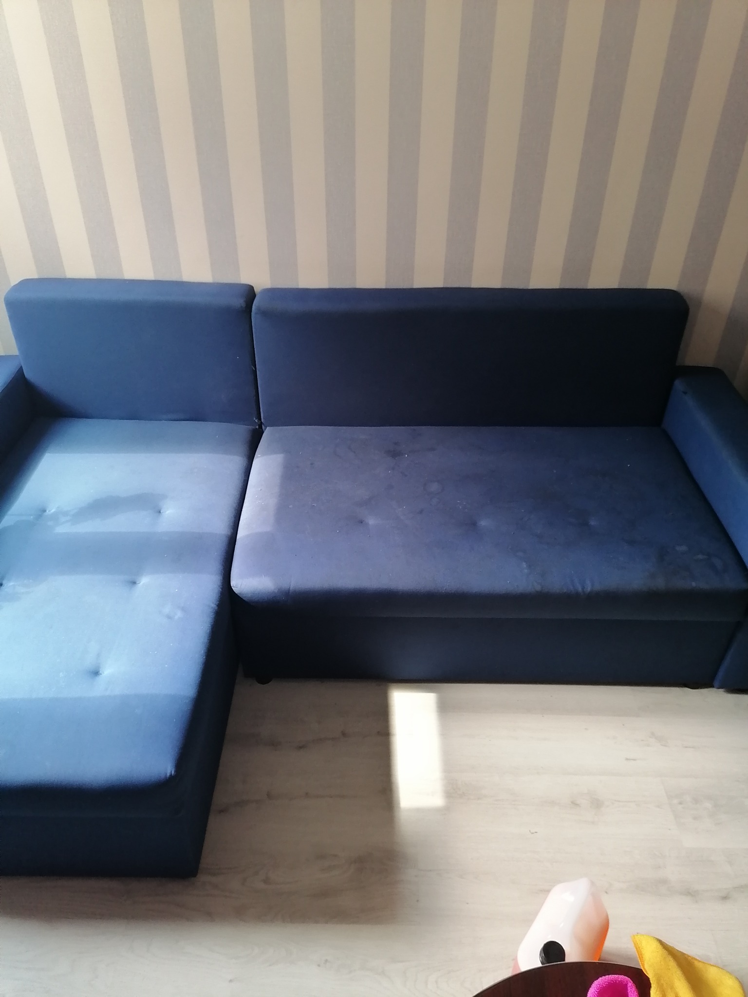 Niebieska kanapa narożna z widocznymi śladami wilgoci po praniu, na podłodze widoczny fragment butelki z płynem do czyszczenia.