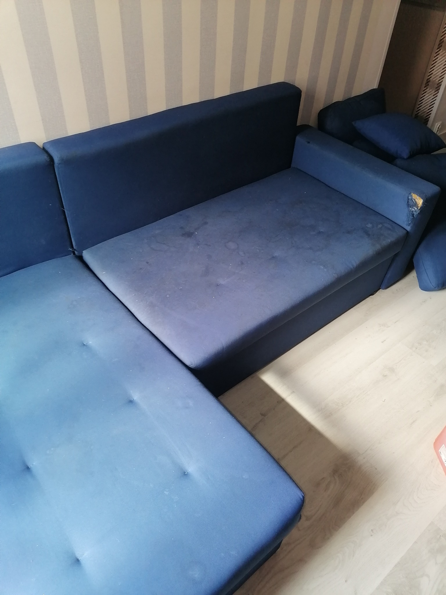 Niebieska, narożna kanapa z widocznymi plamami i uszkodzonym rogiem, stojąca na jasnej podłodze przy ścianie w pasy.