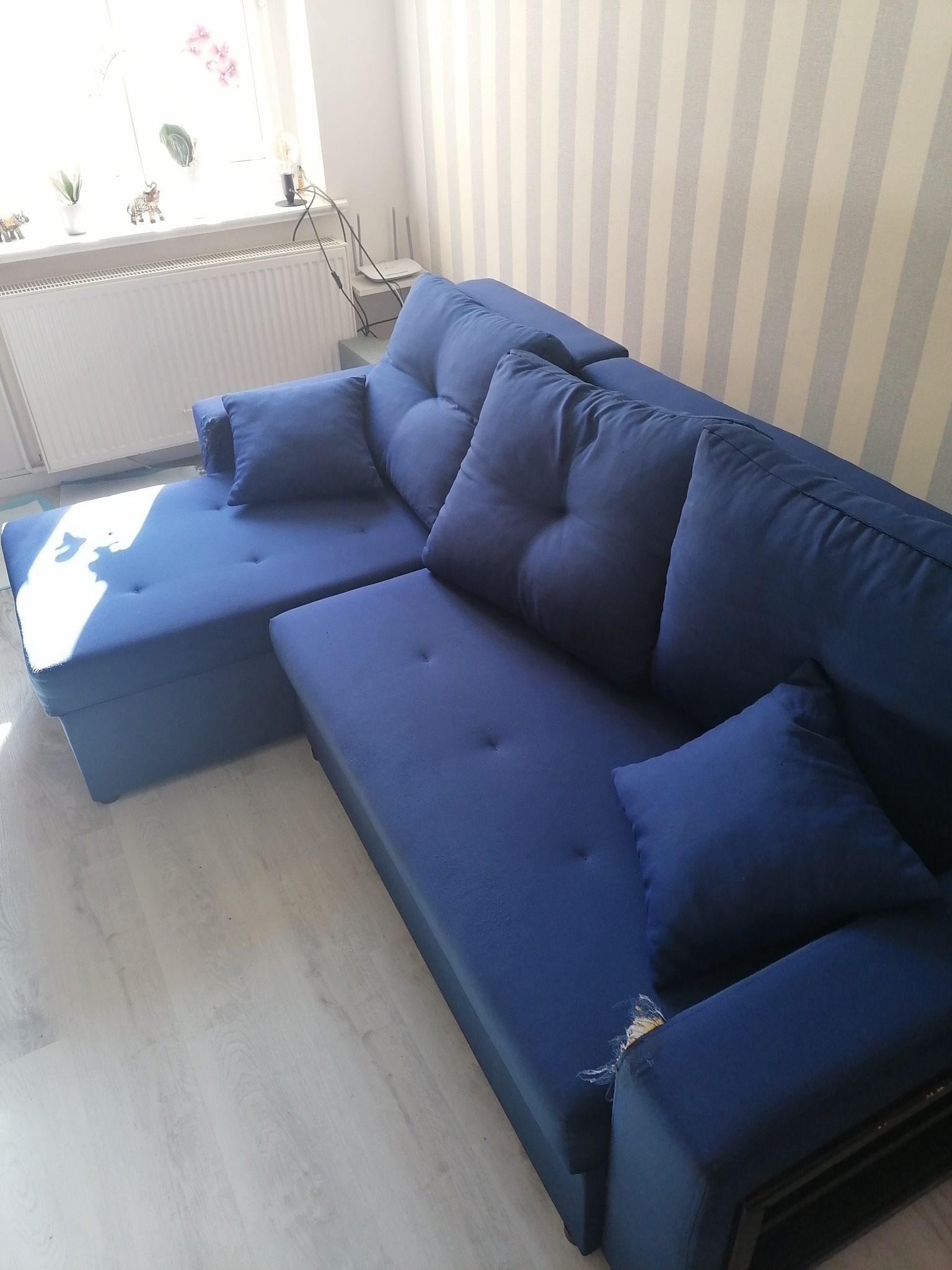 Narożnik tapicerowany w odcieniach granatu z widocznym uszkodzeniem tapicerki na boku, ustawiony w jasnym pomieszczeniu z tapetą w pionowe pasy i grzejnikiem pod oknem.