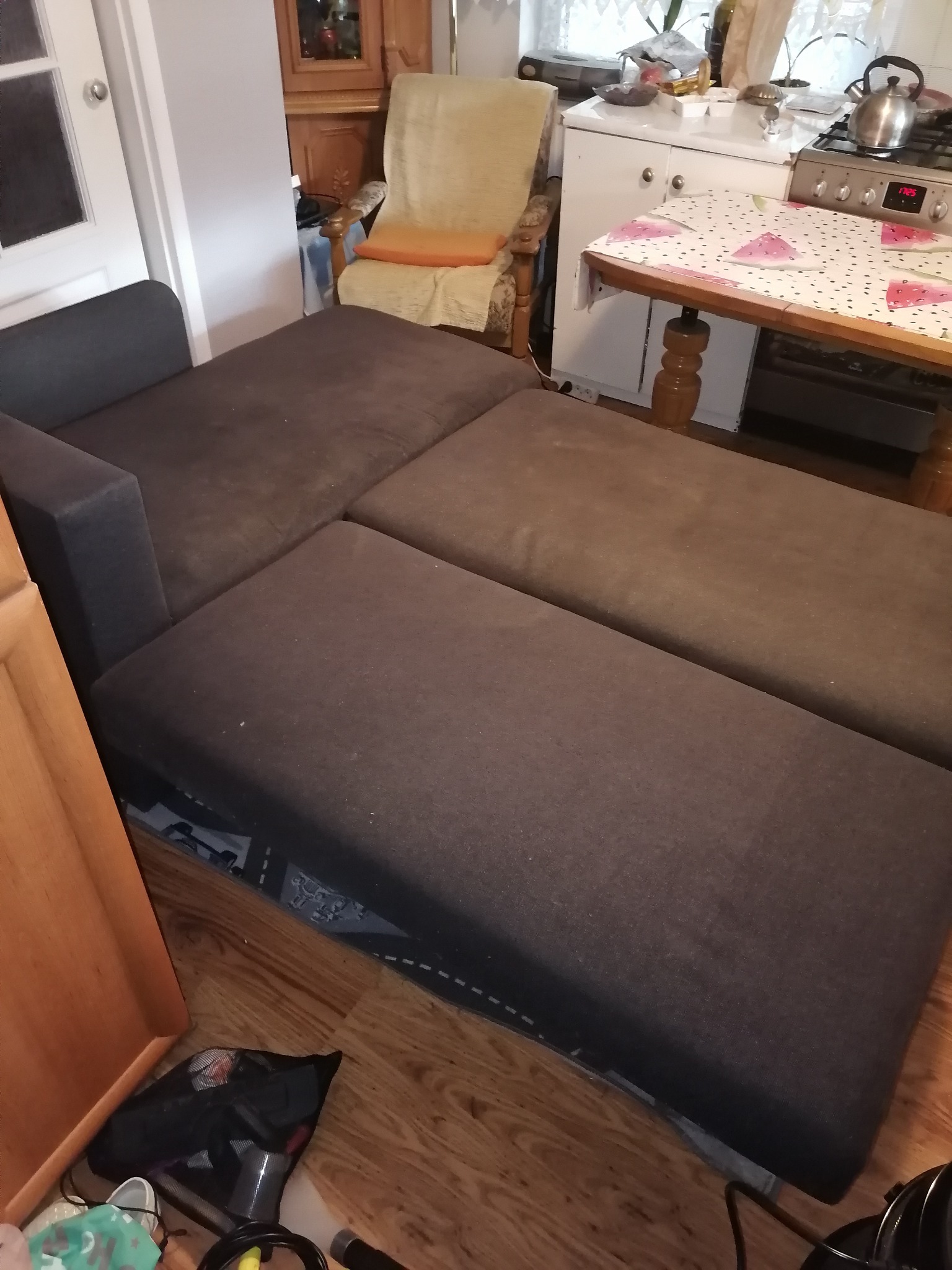 Rozłożona, brązowo-szara kanapa z widocznymi śladami użytkowania, w tle fragment kuchni i inne meble.