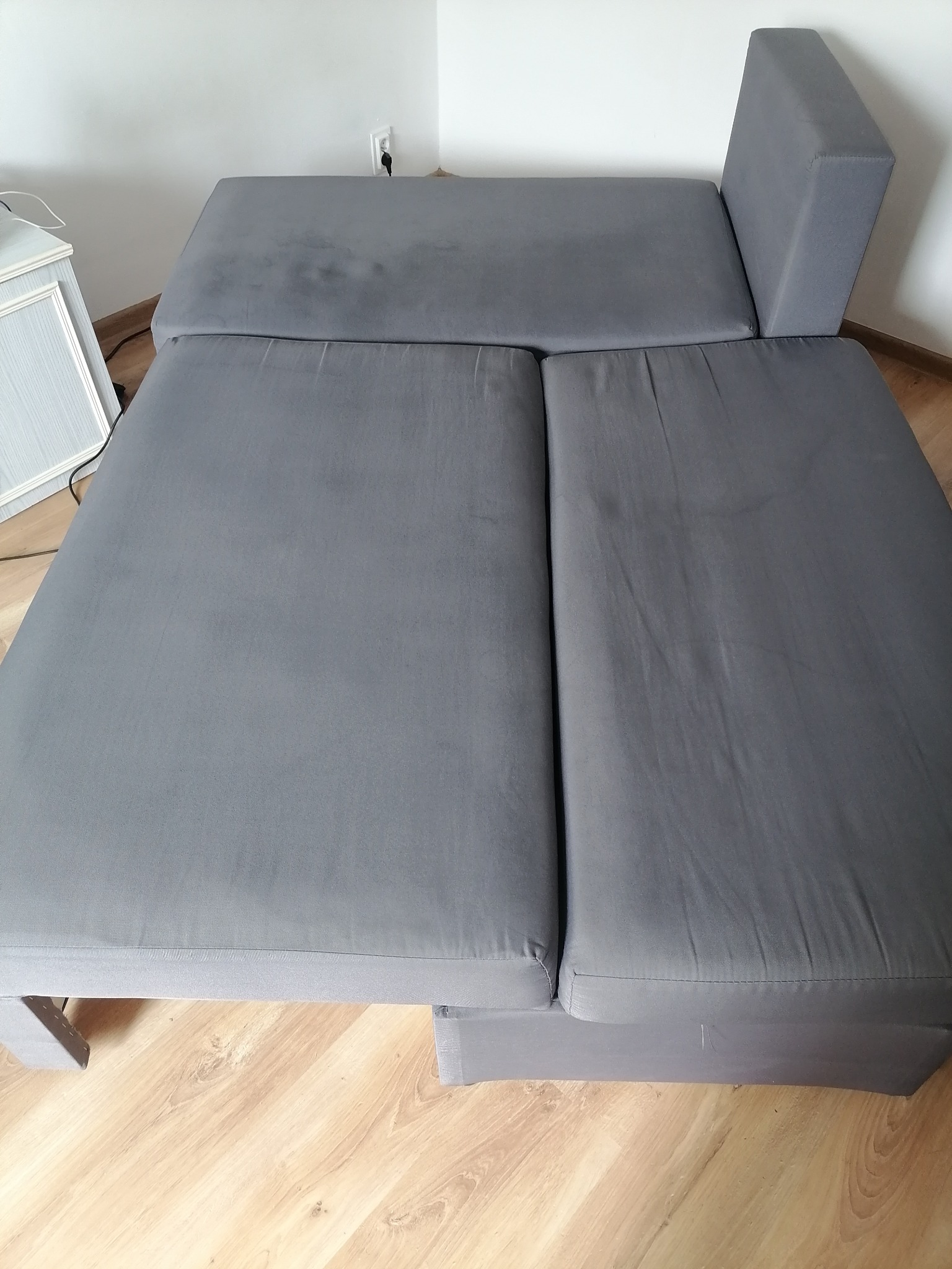 Rozłożona szara kanapa z widocznymi plamami na górnej części, stojąca na panelach podłogowych, z fragmentem białej komody w tle.