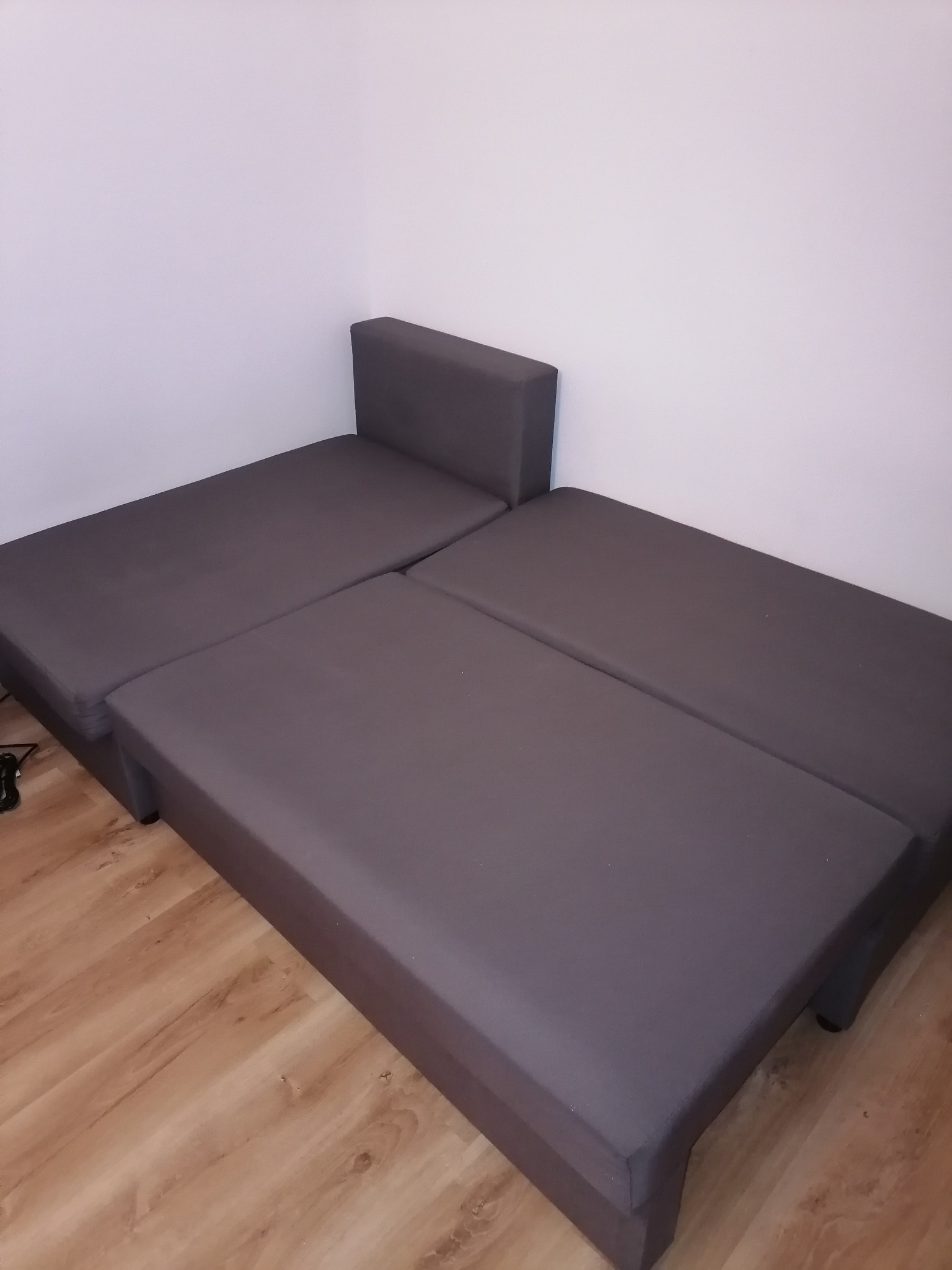 Rozłożona, szara kanapa modułowa z niskim zagłówkiem na drewnianej podłodze, widok z góry w rogu pokoju.