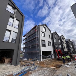 TermoRenowacja - Budowa szeregu domów w trakcie prac wykończeniowych elewacji, widoczne rusztowania, mini koparka i pracownicy w odblaskowych kamizelkach, niebieskie rury na ziemi.