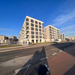 TermoRenowacja - Nowoczesne, wielopiętrowe budynki mieszkalne z balkonami, w jasnej kolorystyce, widziane z poziomu ulicy w słoneczny dzień. W tle dźwig budowlany i inne budynki w budowie. Na pierwszym planie cień...