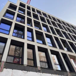 Wielopiętrowy budynek w trakcie prac elewacyjnych, widoczne rusztowania w oknach, dolna część ściany z warstwą izolacyjną i fragmentem niezabezpieczonej powierzchni.