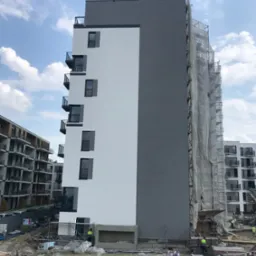 Wielopiętrowy budynek w trakcie ocieplania i malowania elewacji, widoczne rusztowania osłonięte siatką, fragmenty ocieplonej białej i szarej ściany, oraz materiały budowlane i ekipa robotnicza...