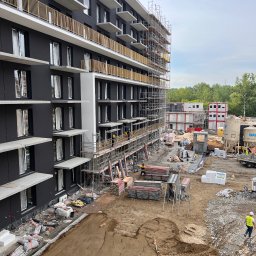 TermoRenowacja - Wielopiętrowy budynek w trakcie prac wykończeniowych elewacji, widoczne rusztowania, materiały budowlane i pracownicy w strojach roboczych, w tle kontenery budowlane i zieleń.