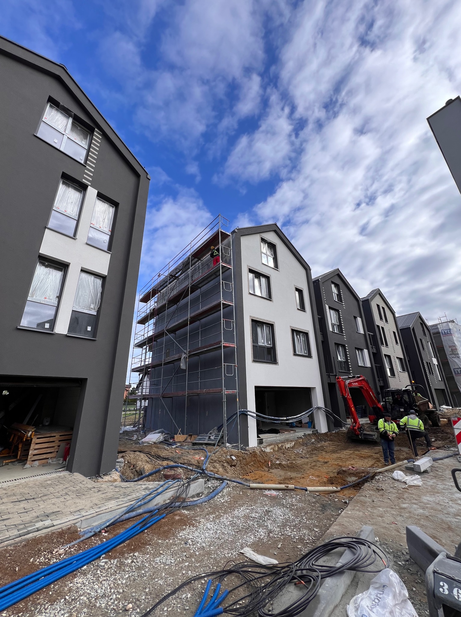 Budowa szeregu domów w trakcie prac wykończeniowych elewacji, widoczne rusztowania, mini koparka i pracownicy w odblaskowych kamizelkach, niebieskie rury na ziemi.