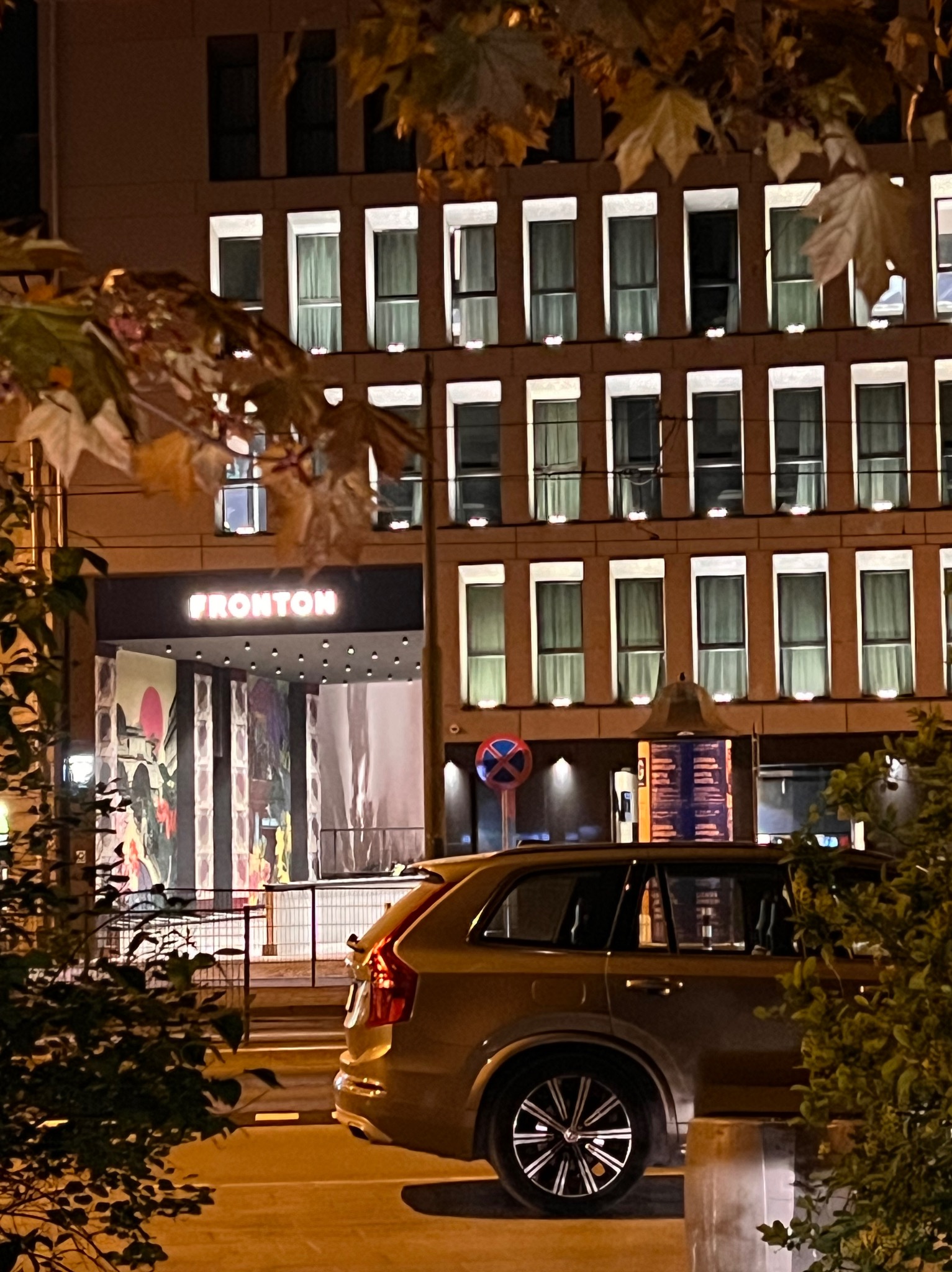 Nocny widok na budynek z wieloma oknami, z nazwą 'FRONTON' nad wejściem, częściowo zasłonięty przez drzewa i zaparkowany samochód.