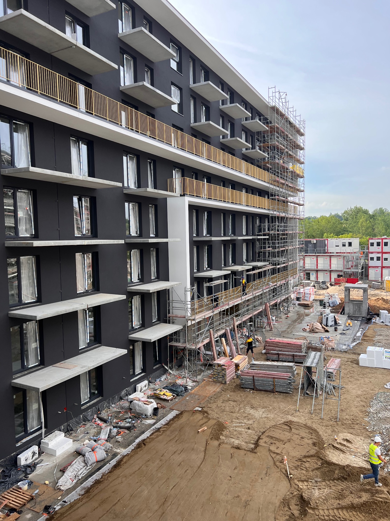 Wielopiętrowy budynek w trakcie ocieplania elewacji, widoczne rusztowania, przygotowane materiały izolacyjne i pracownicy w żółtych kamizelkach.