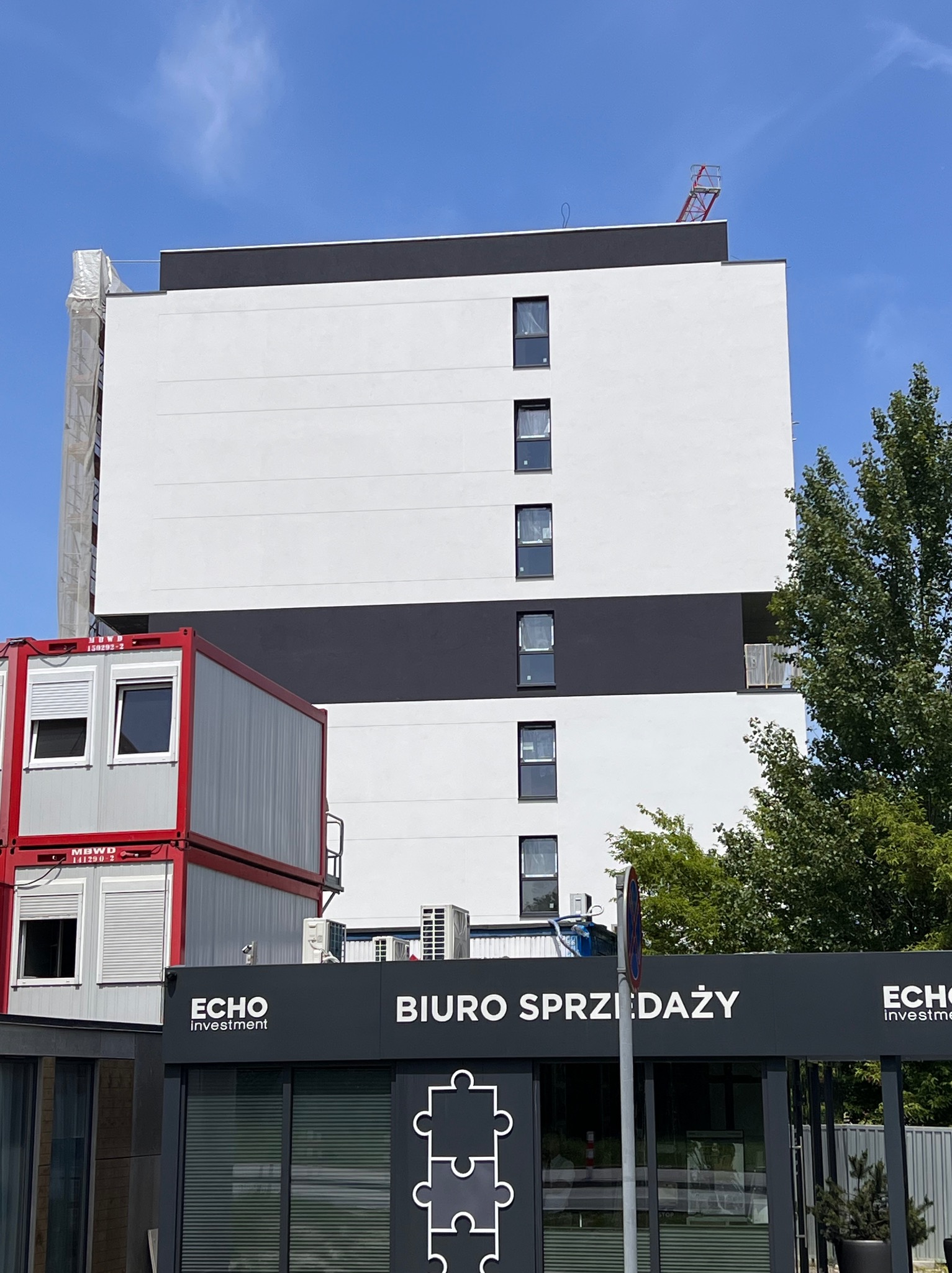 Nowoczesny budynek z białą elewacją i czarnymi pasami, częściowo zakryty przez kontenerowe biuro sprzedaży Echo Investment, z widocznym fragmentem rusztowania i dźwigu na dachu.