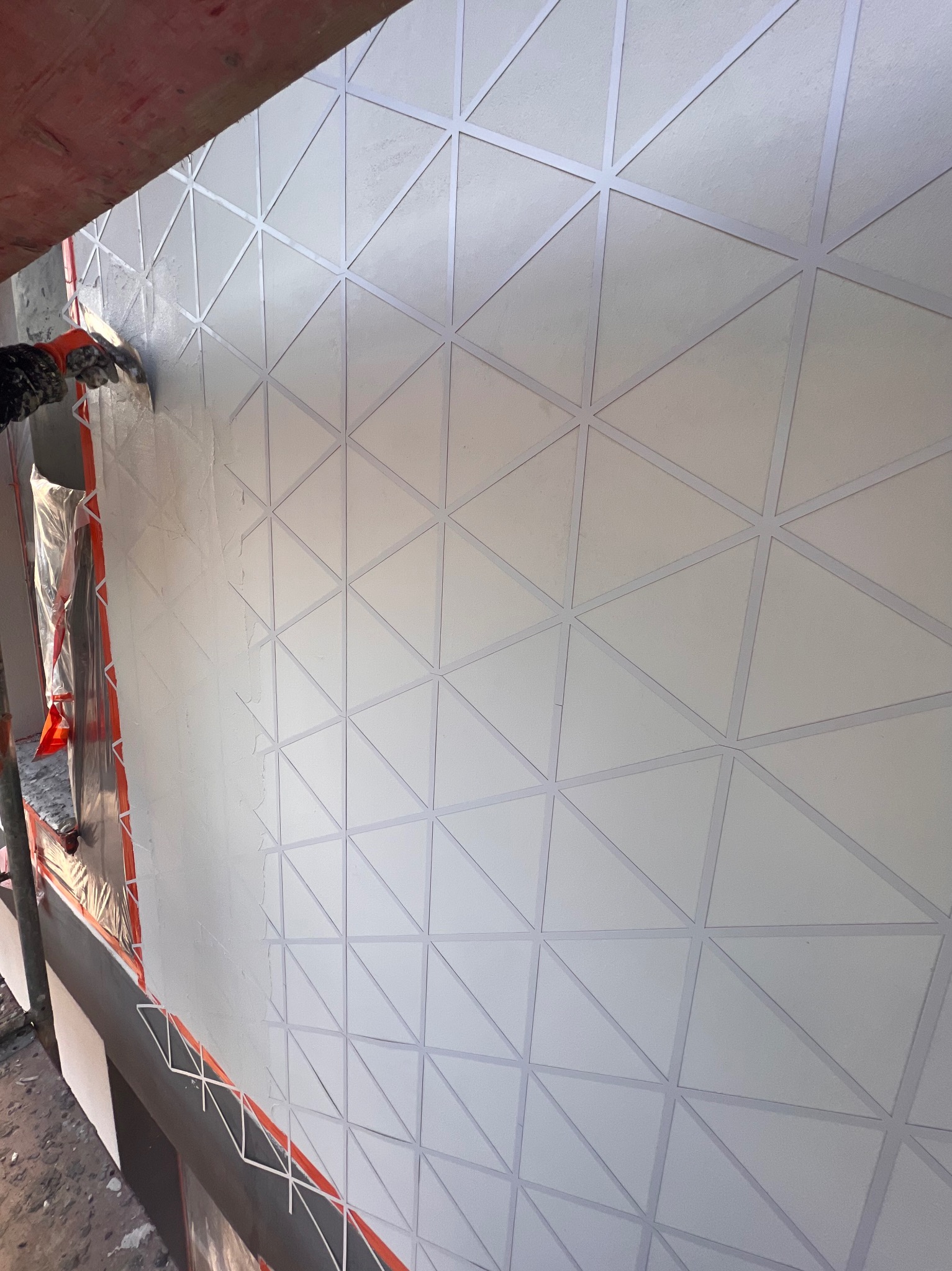 Nakładanie jasnego tynku na elewację z geometrycznym wzorem trójkątów, widoczne rusztowanie i narzędzia pracy.
