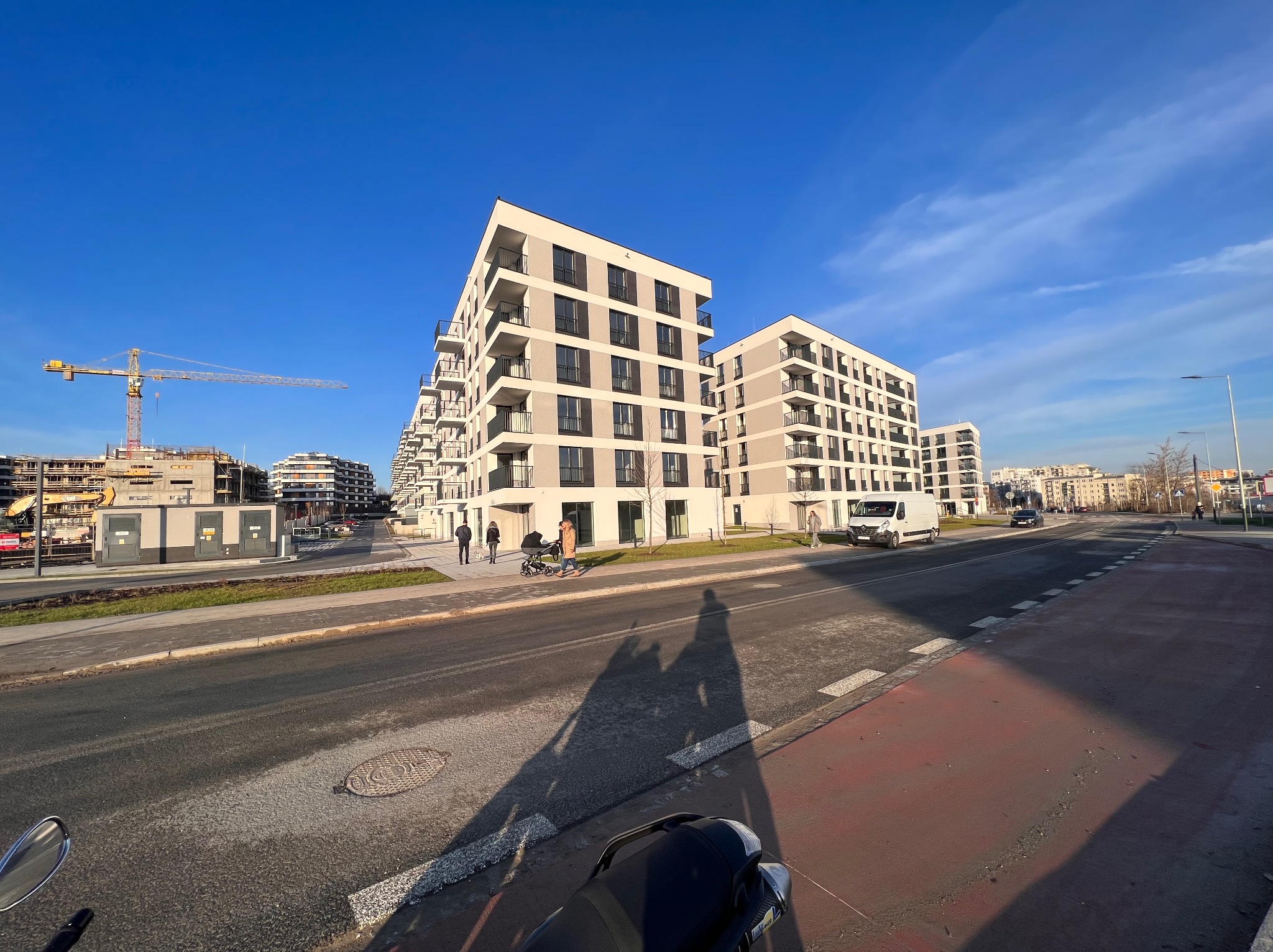 Nowoczesne, wielopiętrowe budynki mieszkalne z balkonami, w jasnej kolorystyce, widziane z poziomu ulicy w słoneczny dzień. W tle dźwig budowlany i inne budynki w budowie. Na pierwszym planie cień...