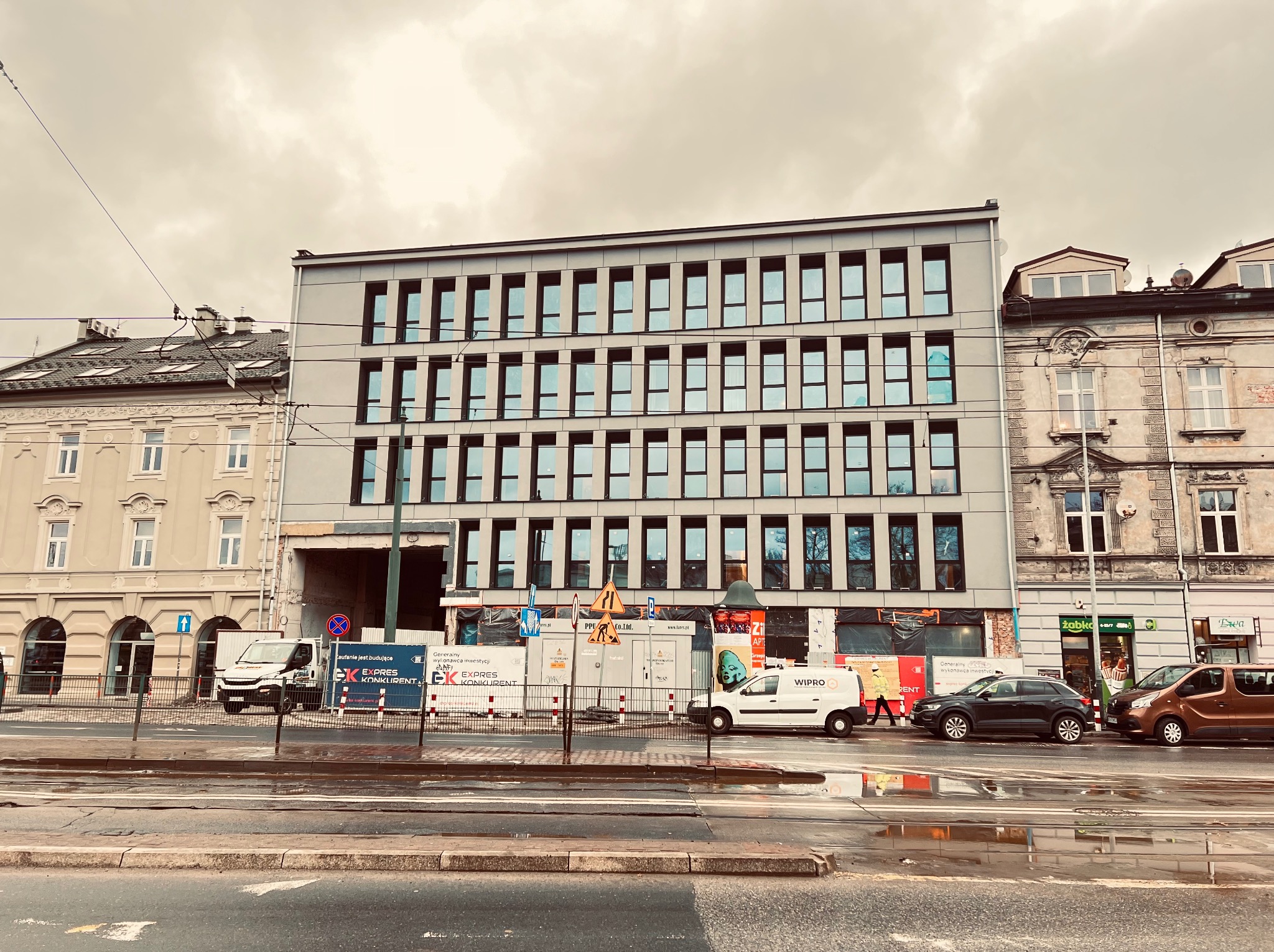 Budynek w trakcie prac elewacyjnych, widoczne rusztowania i materiały budowlane, częściowo odsłonięta ceglana ściana, obok starsza kamienica, mokra ulica po deszczu.