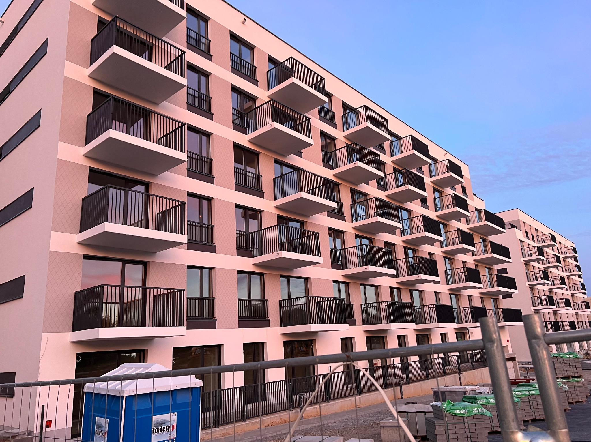 Nowoczesny budynek wielorodzinny z balkonami, różowa elewacja z dekoracyjnym wzorem, widok z dołu, w tle błękitne niebo, elementy placu budowy na pierwszym planie.