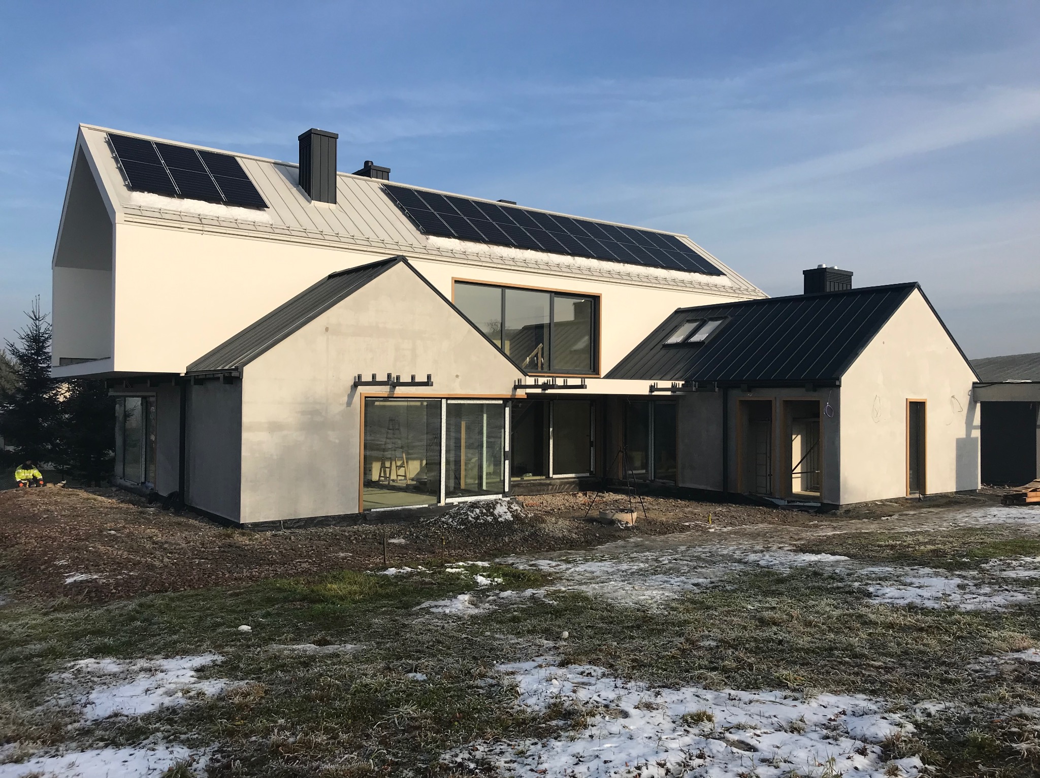 Nowoczesny dom jednorodzinny z panelami słonecznymi na dachu, częściowo otynkowany, z widocznymi oknami i drzwiami tarasowymi, otoczony trawnikiem z resztkami śniegu.
