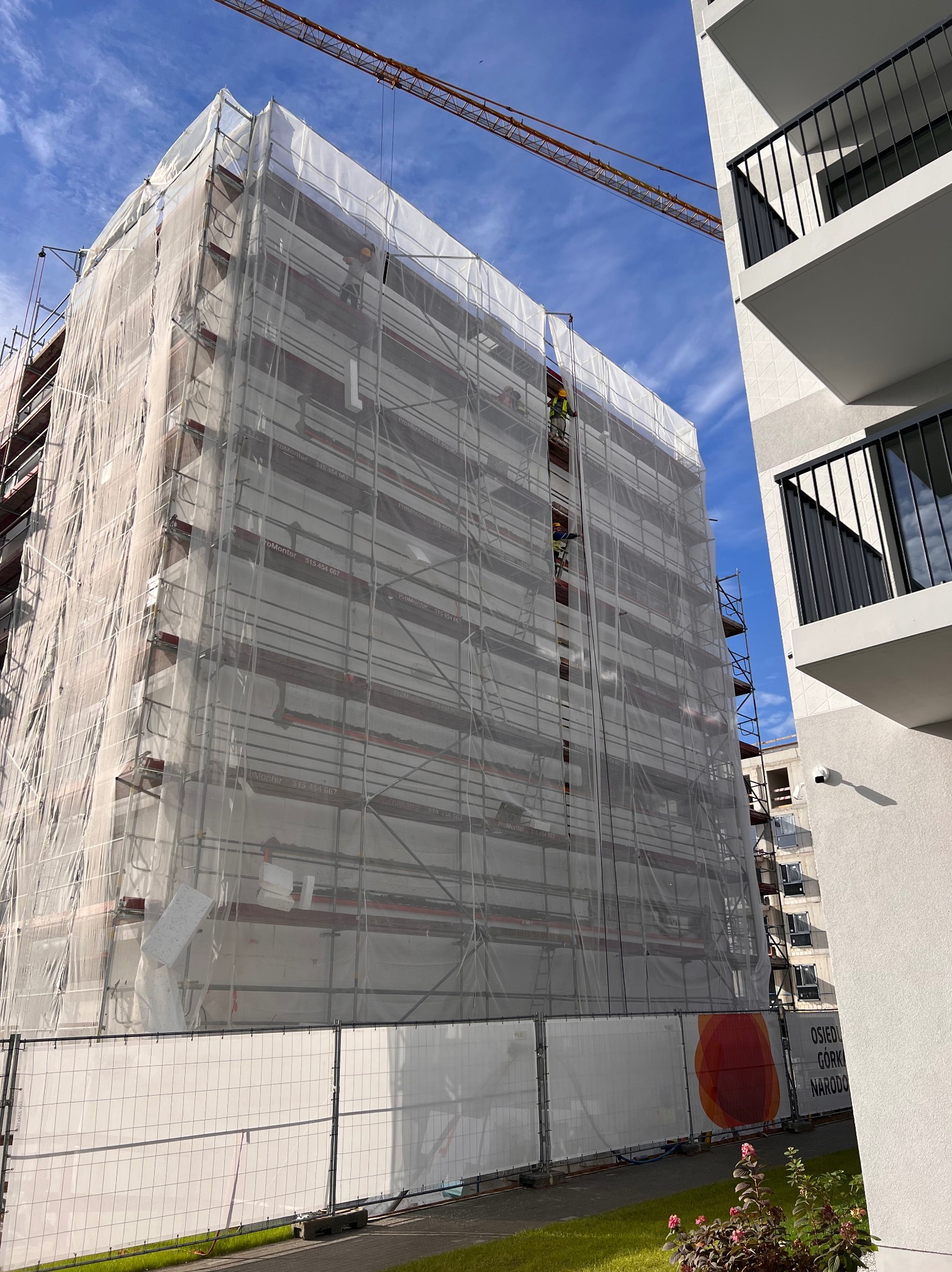 Wielopiętrowy budynek w trakcie prac elewacyjnych, zasłonięty siatką ochronną na rusztowaniach, widoczni pracownicy, w tle błękitne niebo z żurawiem budowlanym, obok fragment nowego budynku...