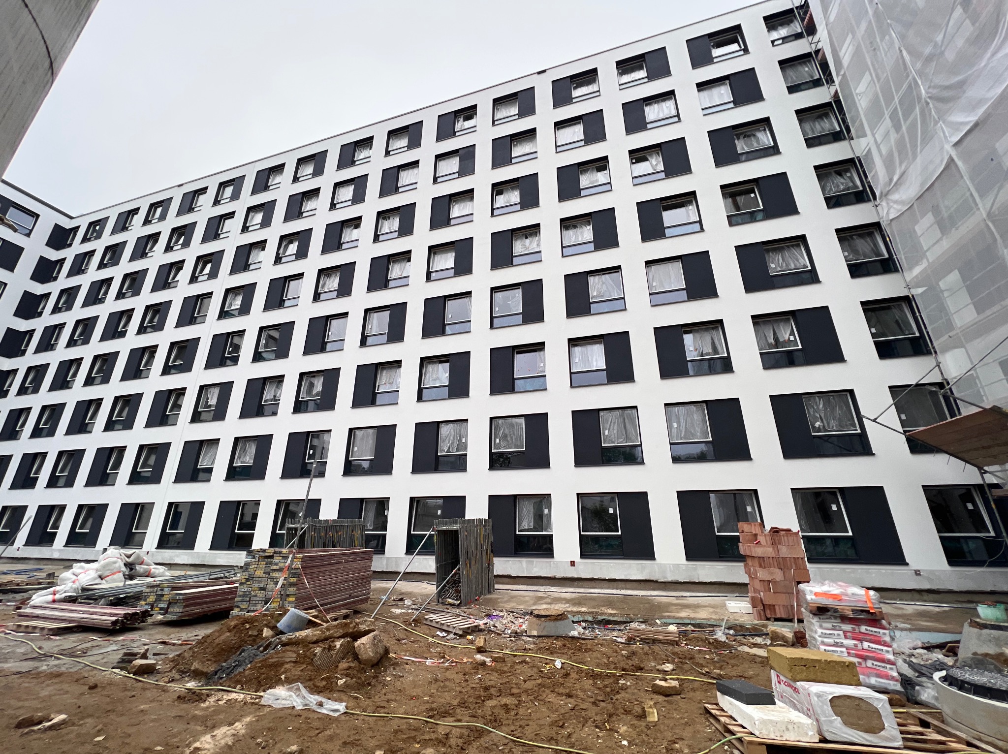 Wielopiętrowy budynek z białą elewacją i czarnymi obramowaniami okien w trakcie prac wykończeniowych, widoczne materiały budowlane i rusztowania.
