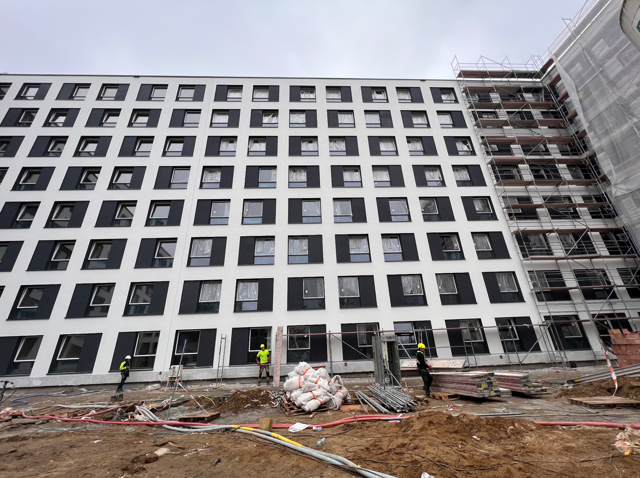 Wielopiętrowy budynek w trakcie prac w Krakowie, widoczne rusztowania, materiały budowlane i pracownicy w odblaskowych kamizelkach. Elewacja częściowo pokryta białym tynkiem, okna zabezpieczone folią.