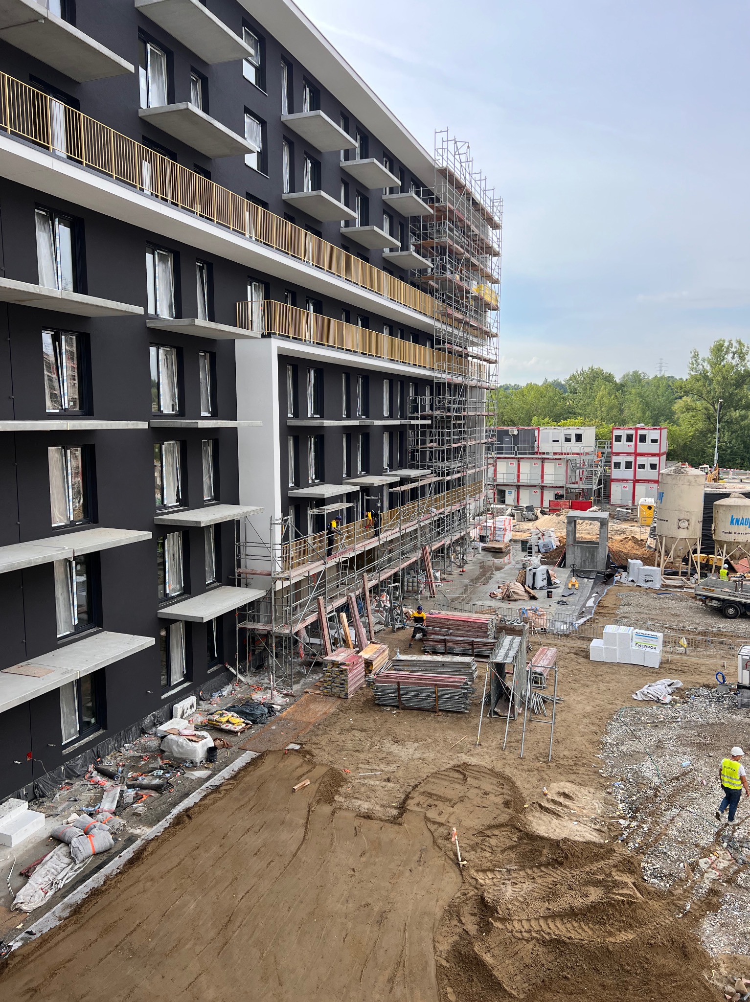 Wielopiętrowy budynek w trakcie prac wykończeniowych elewacji, widoczne rusztowania, materiały budowlane i pracownicy w strojach roboczych, w tle kontenery budowlane i zieleń.