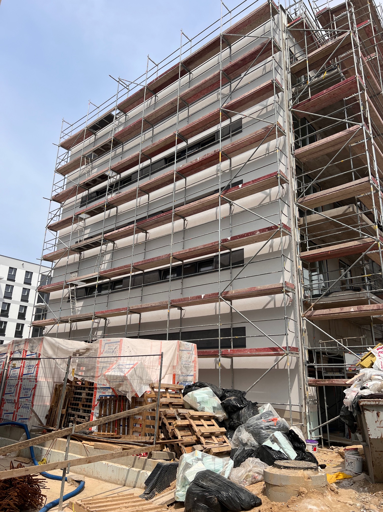 Wielopiętrowy budynek w trakcie prac elewacyjnych, otoczony rusztowaniami, z widocznymi materiałami budowlanymi i workami na śmieci na pierwszym planie.