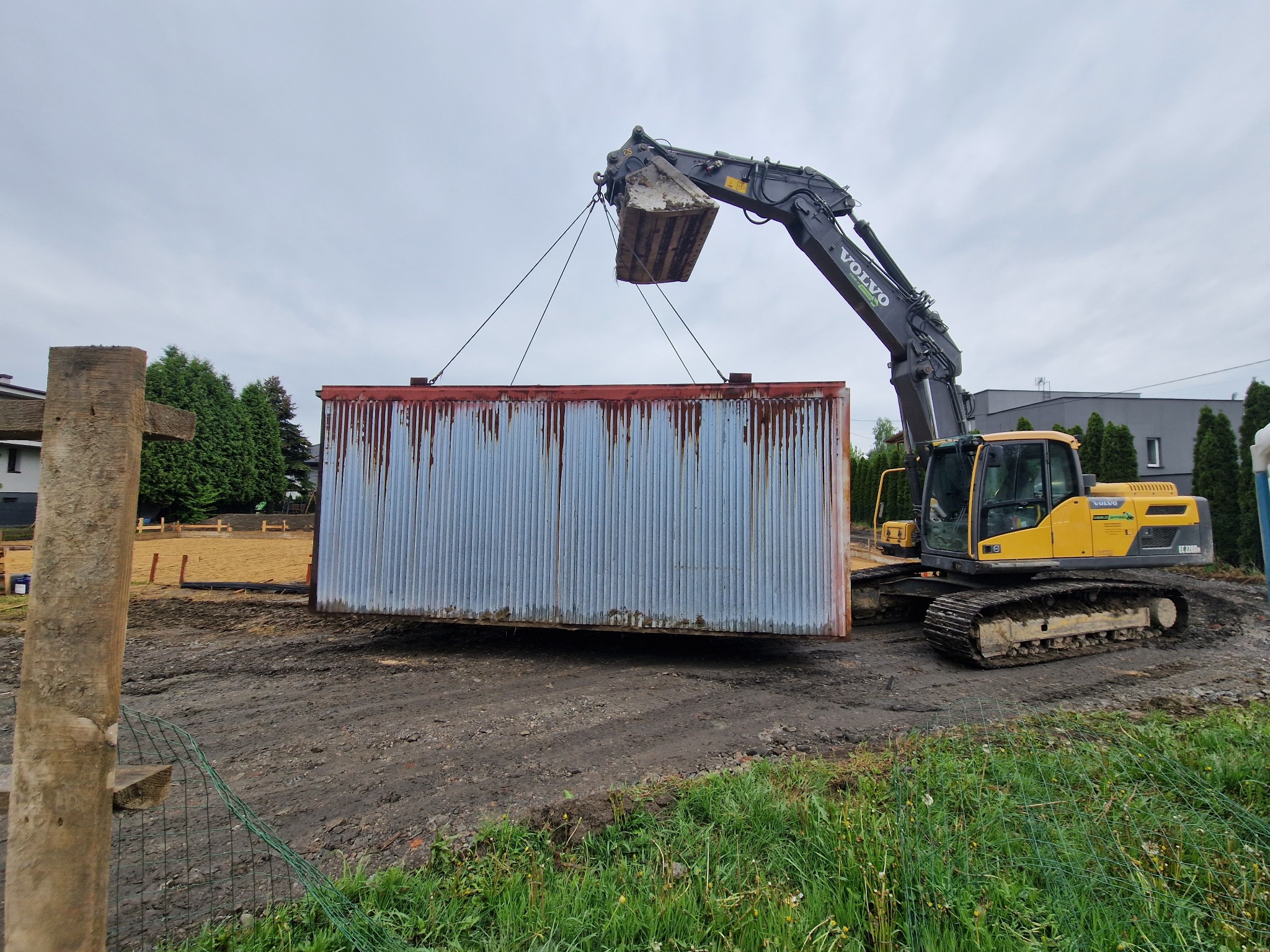 Żółta koparka Volvo podnosi za pomocą lin metalowy, pordzewiały kontener na tle placu budowy i pochmurnego nieba.