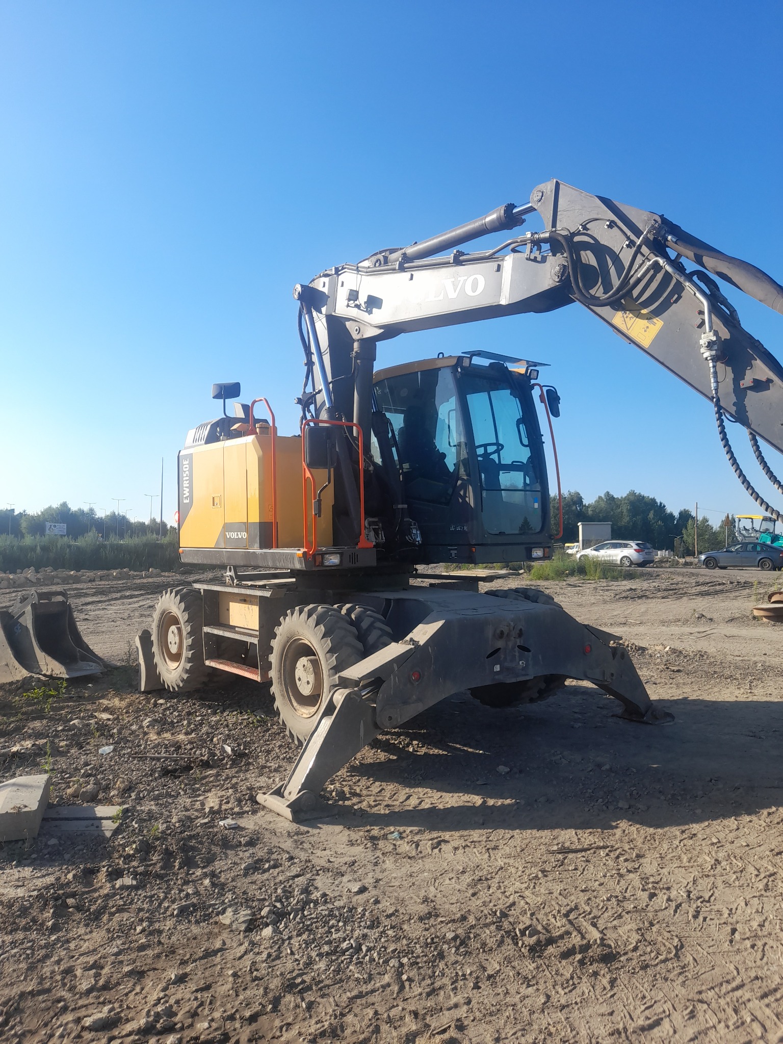 Żółto-szara koparka kołowa Volvo EWR150E stoi na nierównym, piaszczystym terenie budowy w słoneczny dzień. Widoczne stabilizatory, ramię koparki i łyżka obok. W tle zaparkowane samochody.