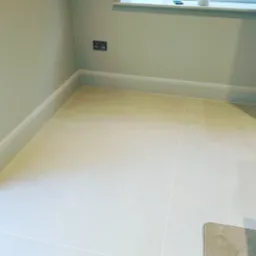 Jasne, kwadratowe płytki podłogowe w odcieniach beżu, ułożone w pomieszczeniu w Jeleniej Górze, z widoczną listwą przypodłogową w kolorze ściany i gniazdkiem elektrycznym.