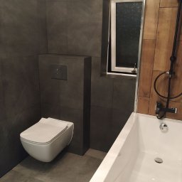 Prestige Tiling - Nowoczesna łazienka z szarymi płytkami, podwieszaną toaletą, drewnianymi panelami przy wannie i czarną armaturą, okno uchylne w tle.