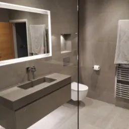 Nowoczesna łazienka w szarościach z podświetlanym lustrem, wiszącą toaletą i grzejnikiem drabinkowym z ręcznikiem.