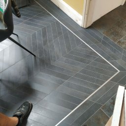 Prestige Tiling - Ułożona w jodełkę, grafitowa podłoga z płytek ceramicznych, oddzielona listwą od starej podłogi, widoczne fragmenty krzesła i roboczych materiałów.