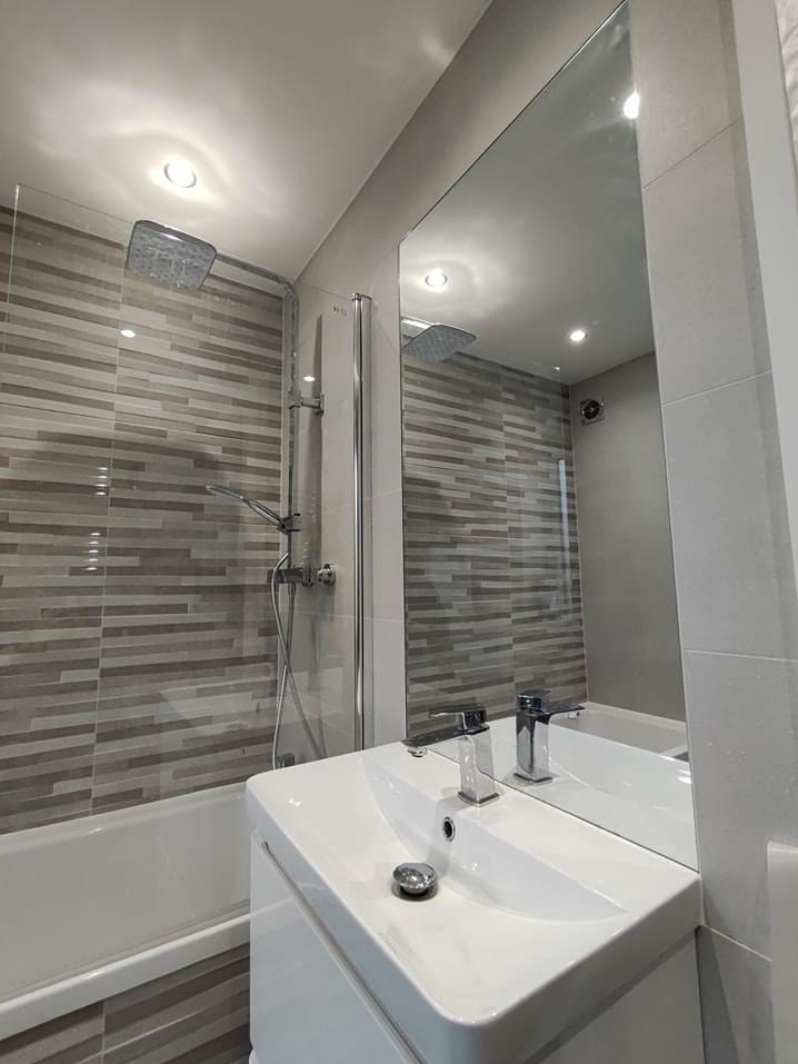 Nowoczesna łazienka z poziomymi płytkami w odcieniach szarości, kabiną prysznicową typu walk-in i białą umywalką z chromowaną baterią, odbijającą się w dużym lustrze.