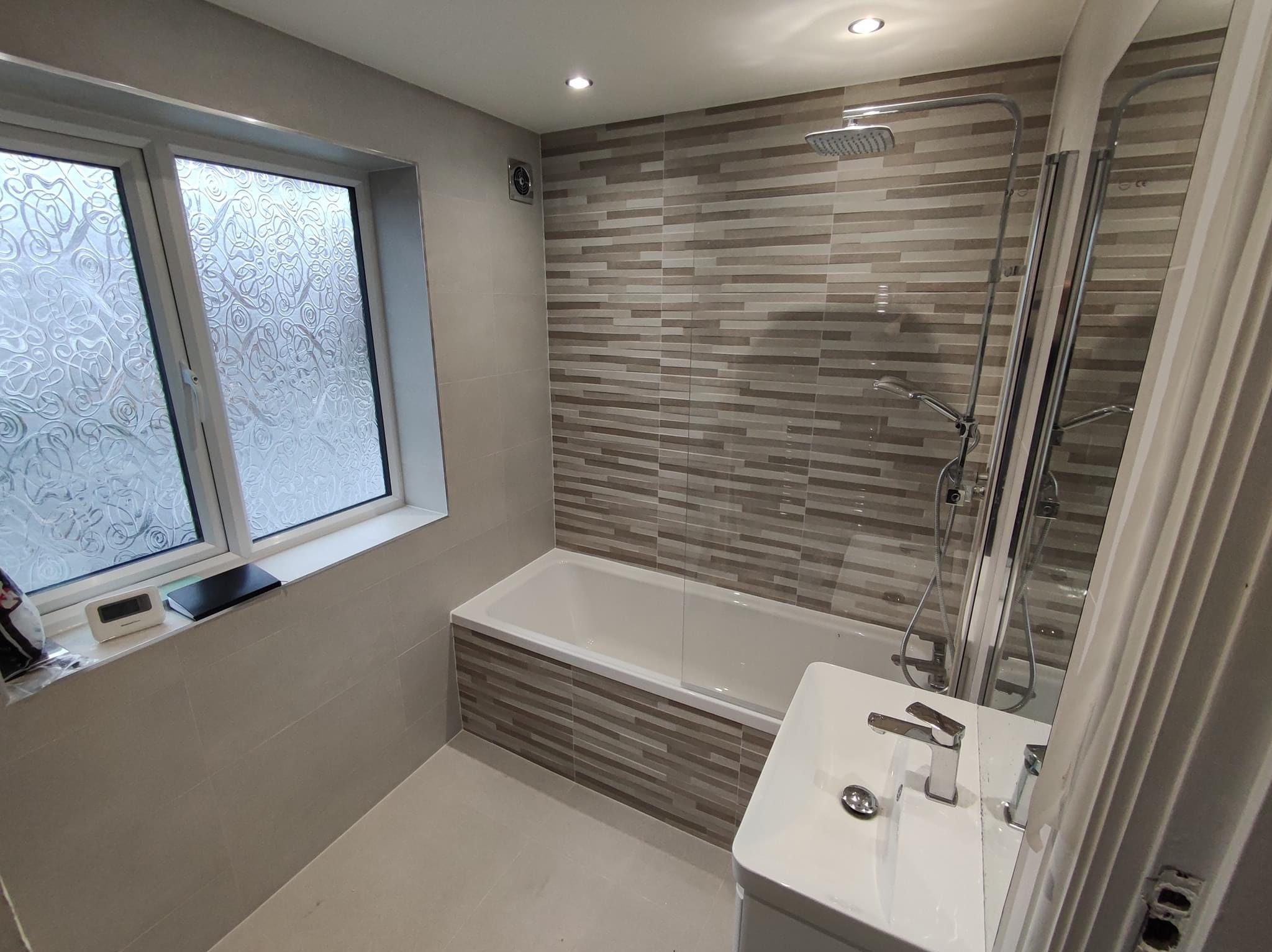 Nowoczesna łazienka z wanną obudowaną płytkami imitującymi drewno, szklanym parawanem nawannowym, umywalką nablatową i oknem z ozdobną szybą.