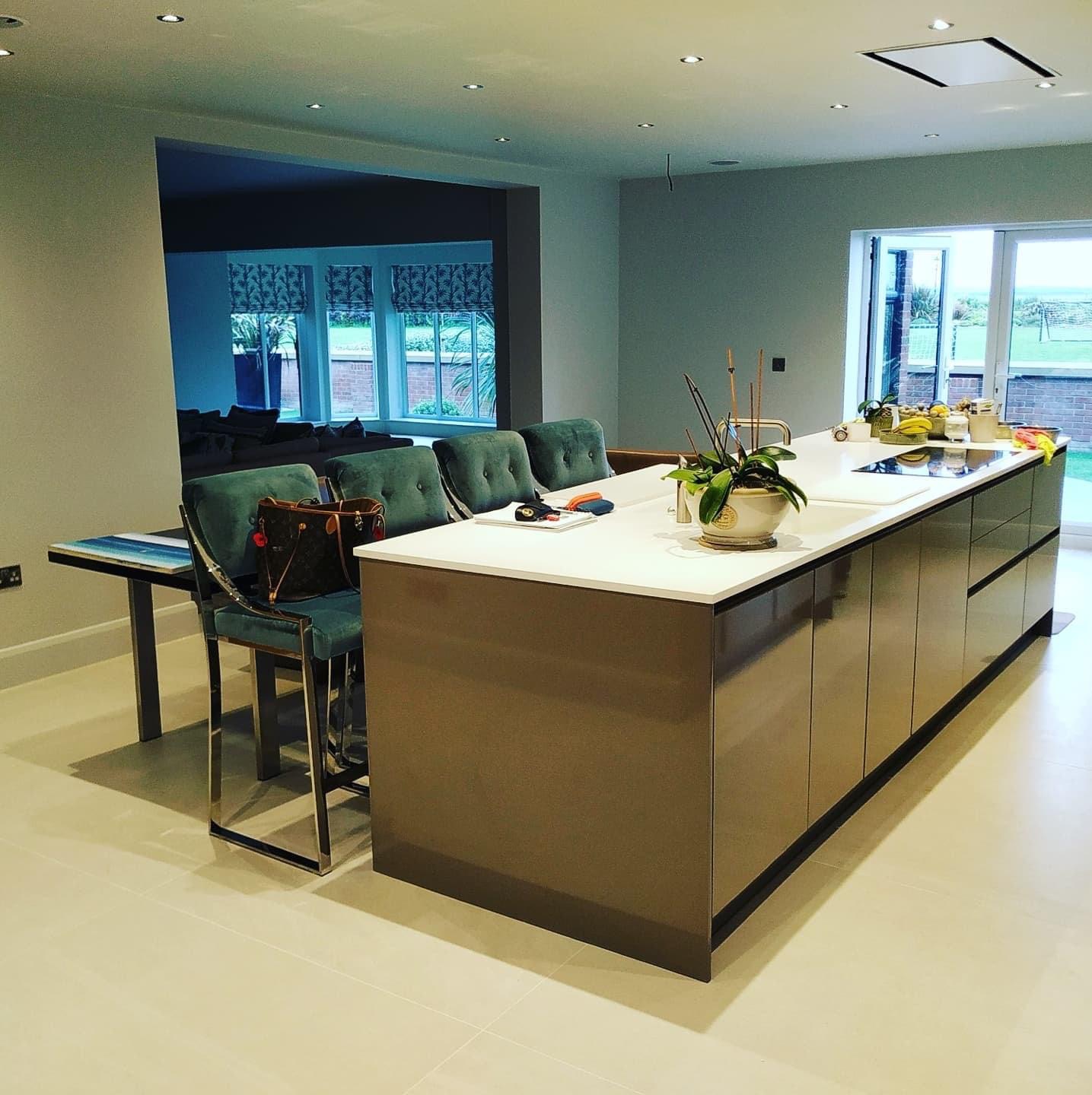 Nowoczesna kuchnia z wyspą w odcieniach brązu, białym blatem, zielonymi krzesłami barowymi i widokiem na salon przez szerokie przejście.