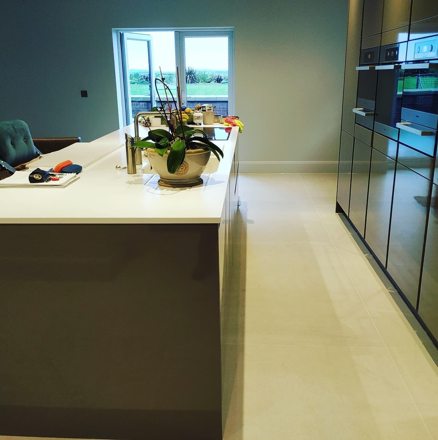 Nowoczesna kuchnia z białym blatem, ciemnymi szafkami i beżową podłogą. Na blacie stoi doniczka z kwiatem, a w tle widać otwarte drzwi balkonowe z widokiem na zieleń.