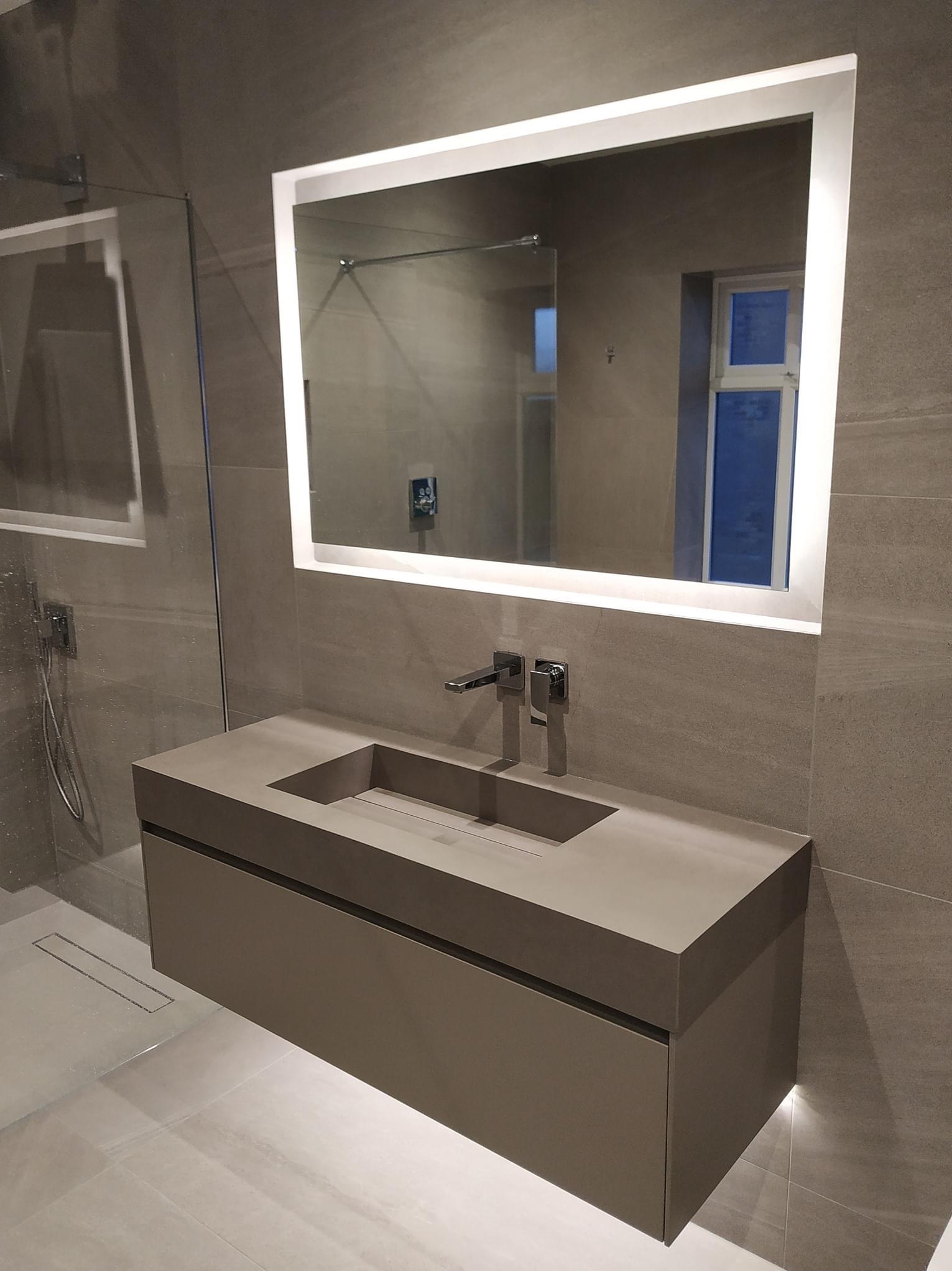 Nowoczesna łazienka z szarą, podwieszaną szafką z umywalką o geometrycznym kształcie i podświetlanym lustrem LED, wykończona płytkami w odcieniach szarości, z widoczną kabiną prysznicową.