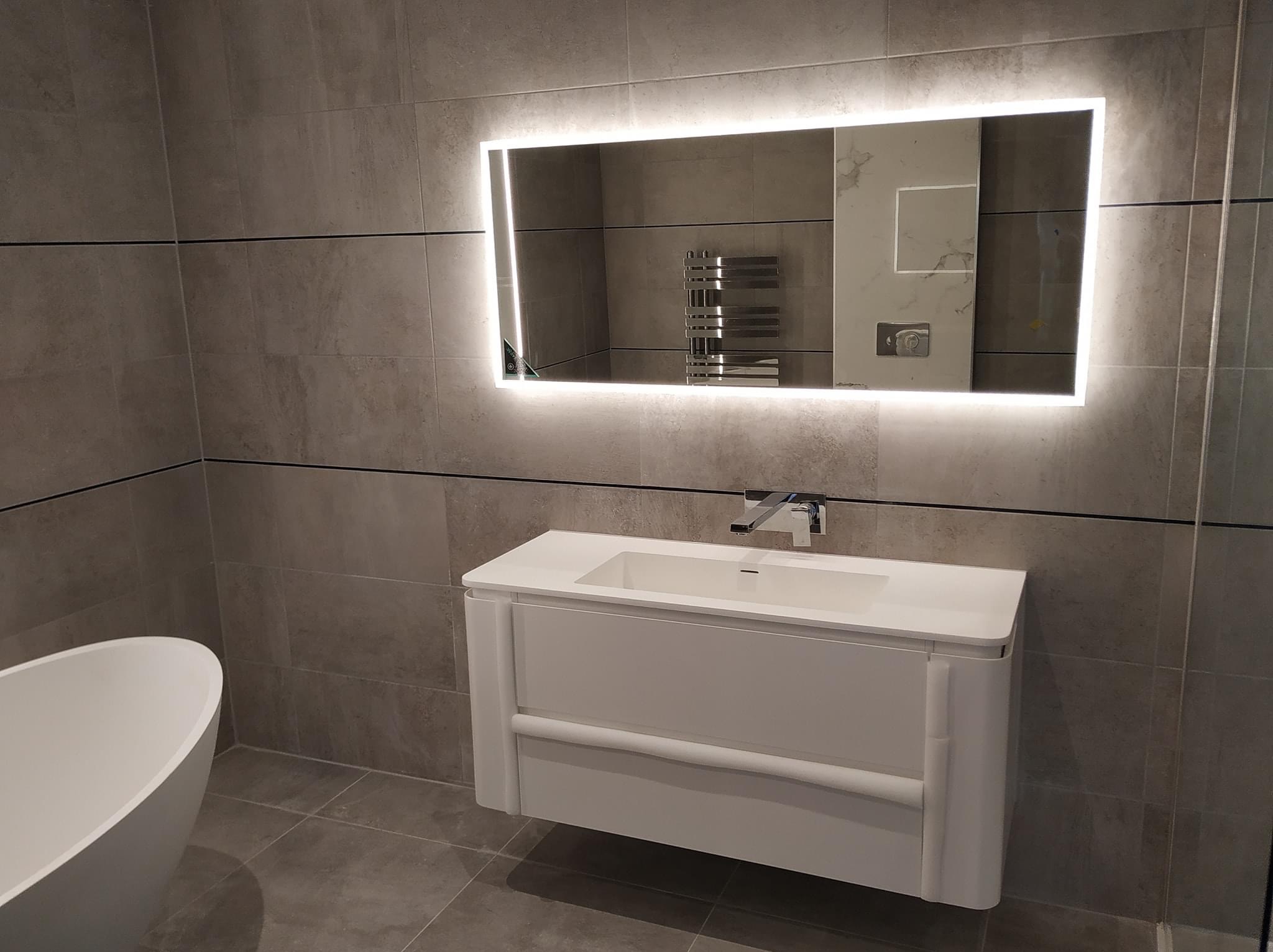 Nowoczesna łazienka z szarymi płytkami, podświetlanym lustrem, białą szafką z umywalką i fragmentem wanny wolnostojącej.