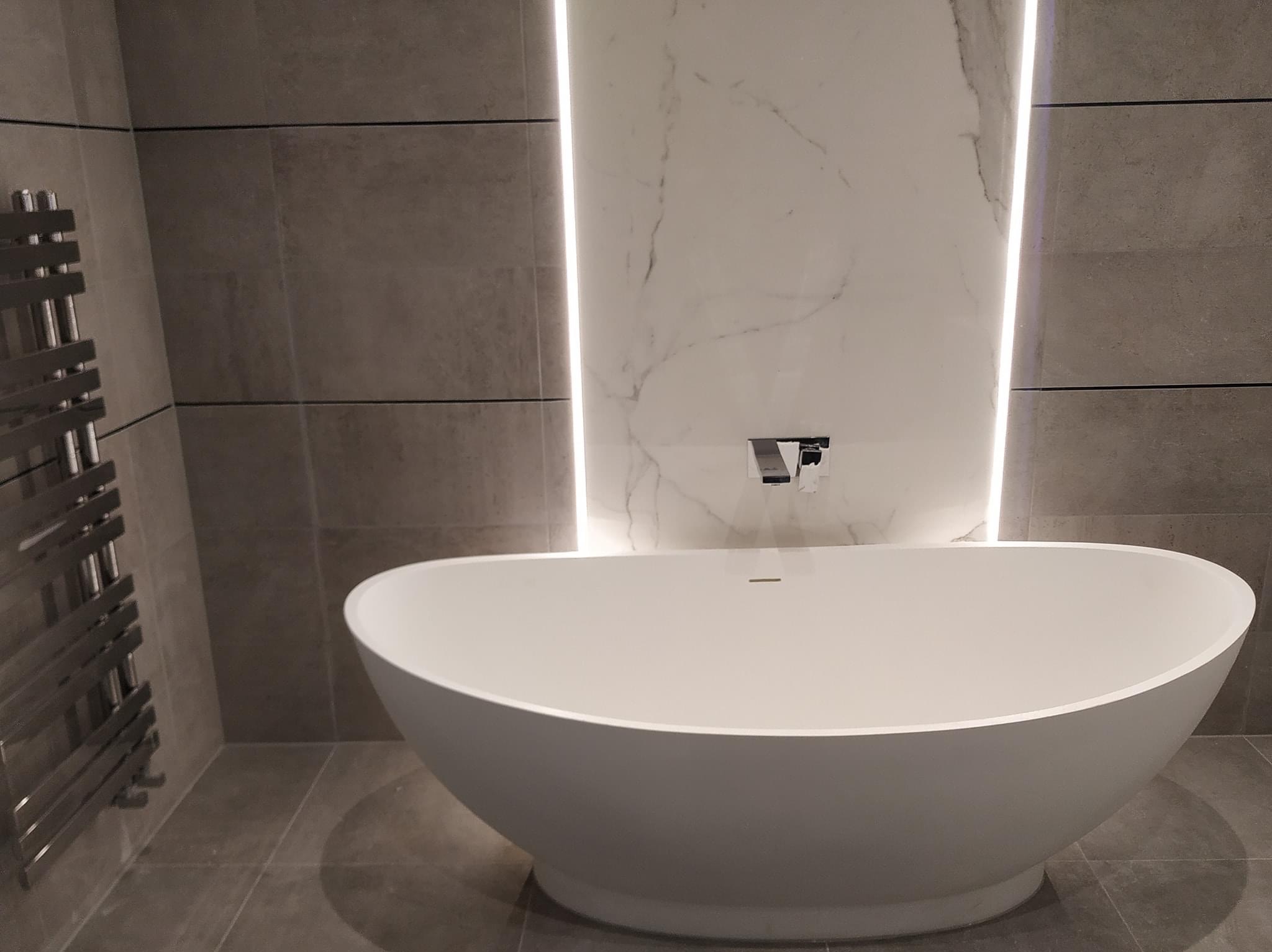 Wolnostojąca, owalna wanna z chromowaną baterią, podświetlona pionowymi pasami LED na tle płytek imitujących marmur i szarych płytek ściennych oraz grzejnik drabinkowy.
