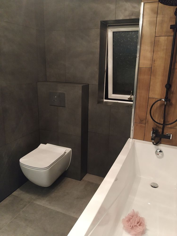 Nowoczesna łazienka z szarymi płytkami, podwieszaną toaletą, drewnianymi panelami przy wannie i czarną armaturą, okno uchylne w tle.