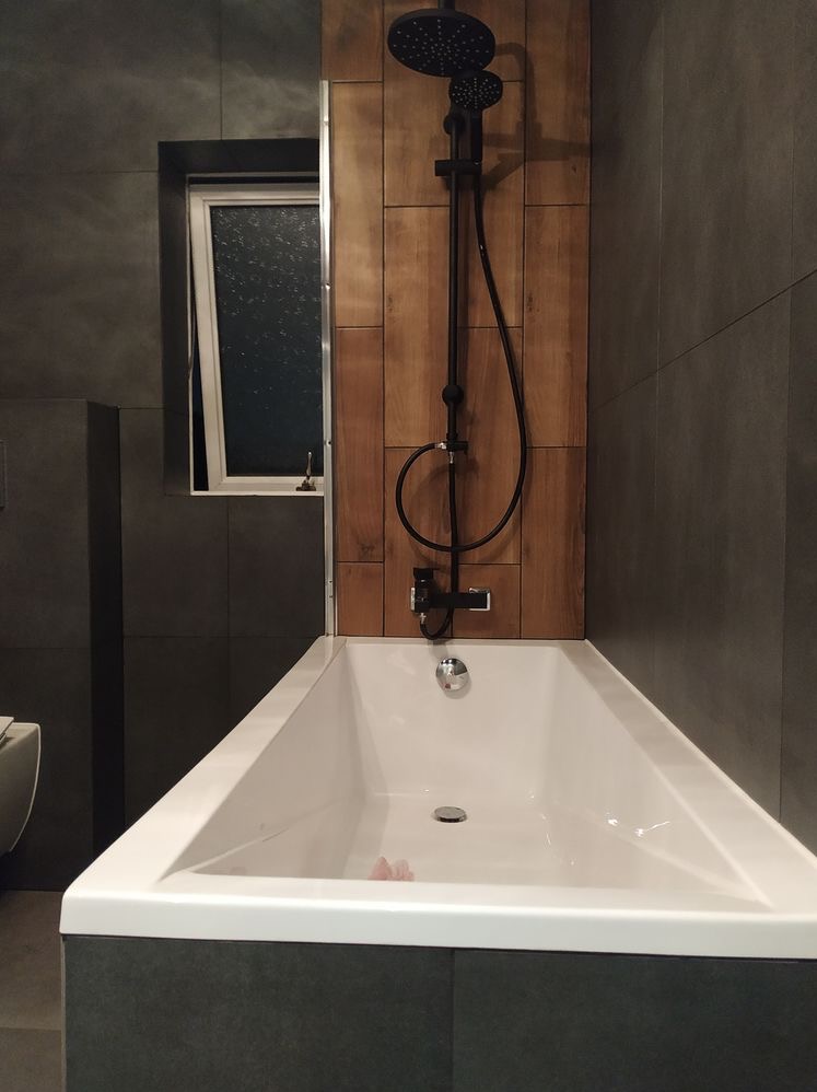 Nowoczesna łazienka z wanną w zabudowie, czarna armatura, drewnopodobne panele na ścianie i uchylne okno.