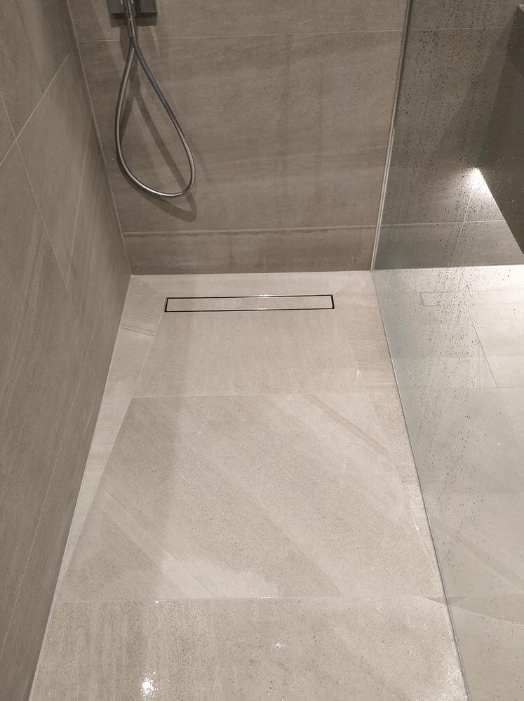 Nowoczesny prysznic walk-in z odpływem liniowym i szarymi płytkami imitującymi kamień, widoczne krople wody na szklanej ściance.