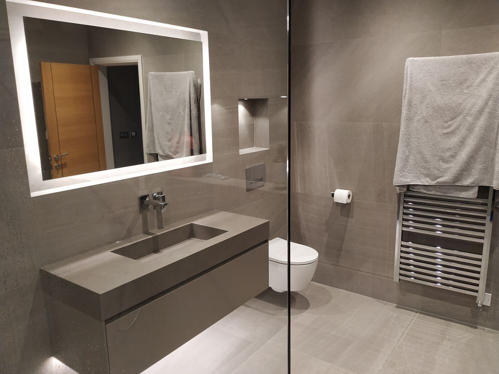 Nowoczesna łazienka w szarościach z podświetlanym lustrem, wiszącą toaletą i grzejnikiem drabinkowym z ręcznikiem.